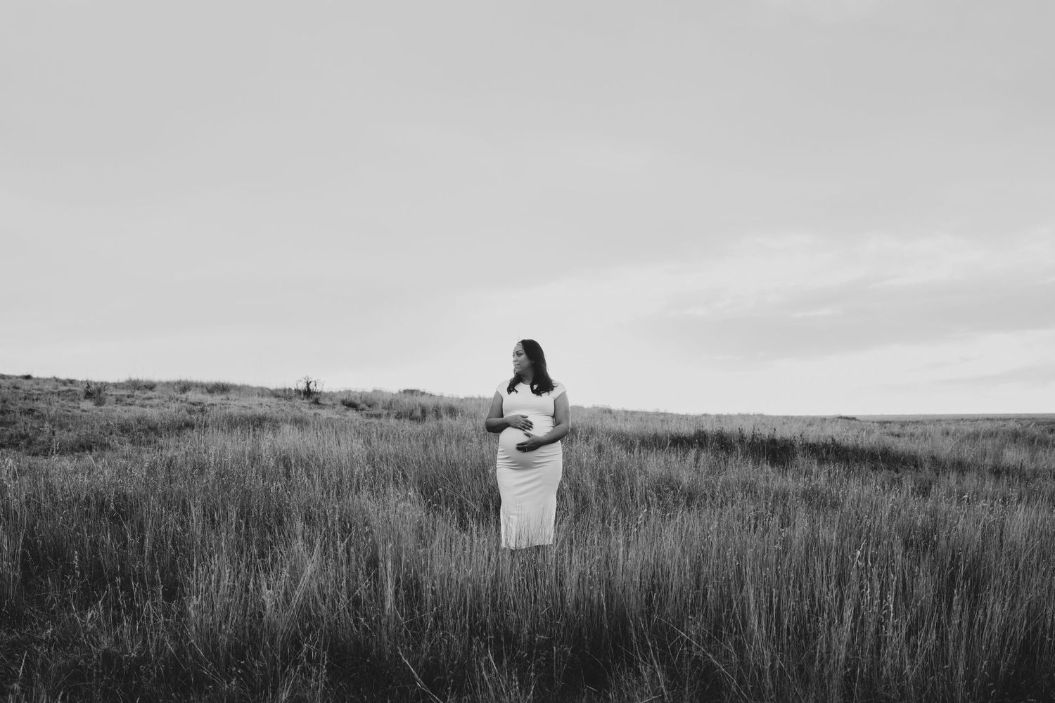 Black and white photo of pregnant woman standing alone in a vast open grass field.