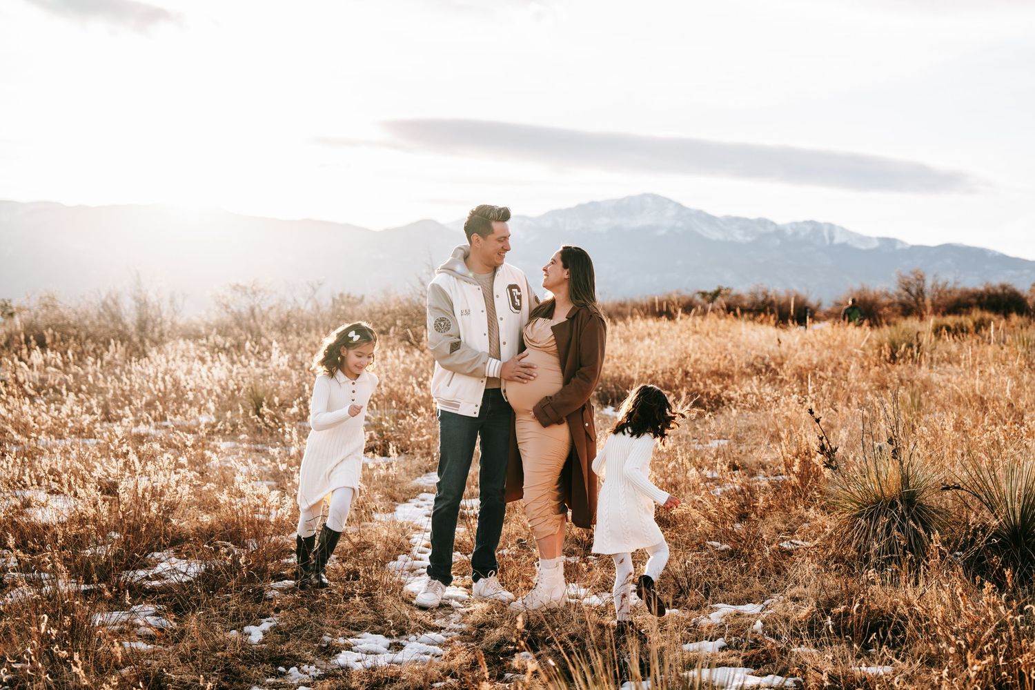 Family of four with pregnant mother standing in golden field with snow patches and mountains at sunset.