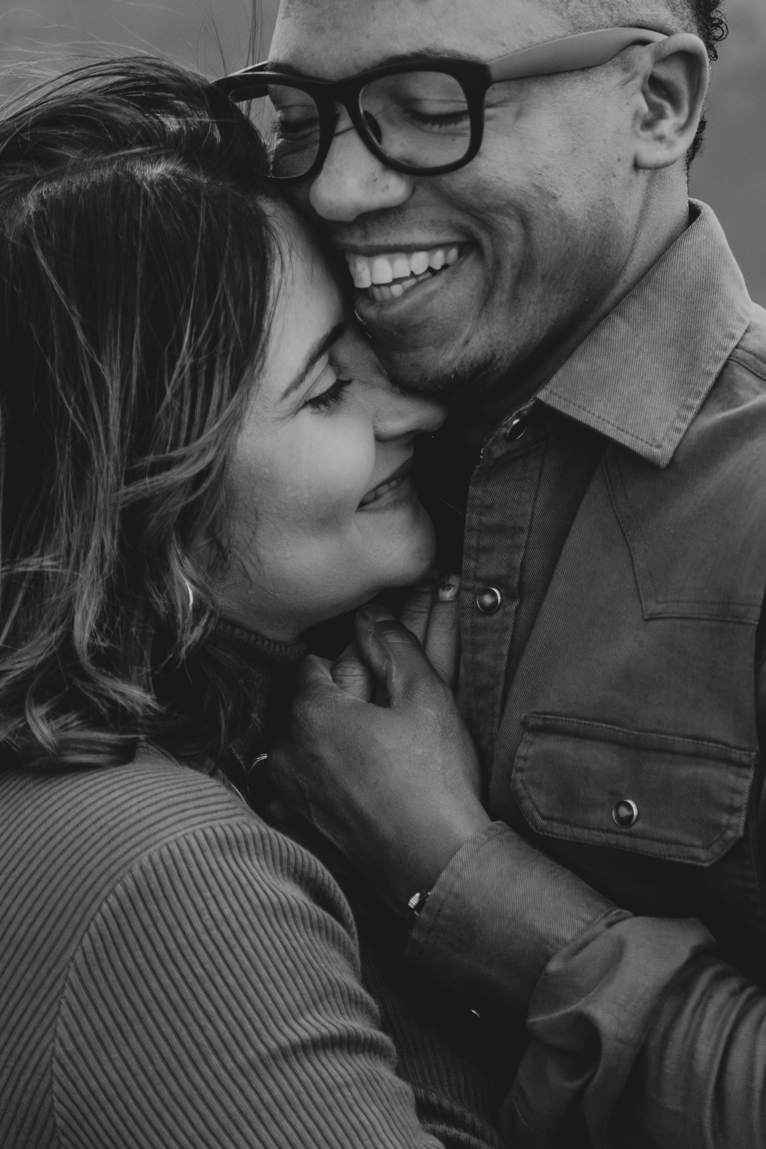 Black and white close-up of a smiling couple embracing, man wearing glasses laughing joyfully.
