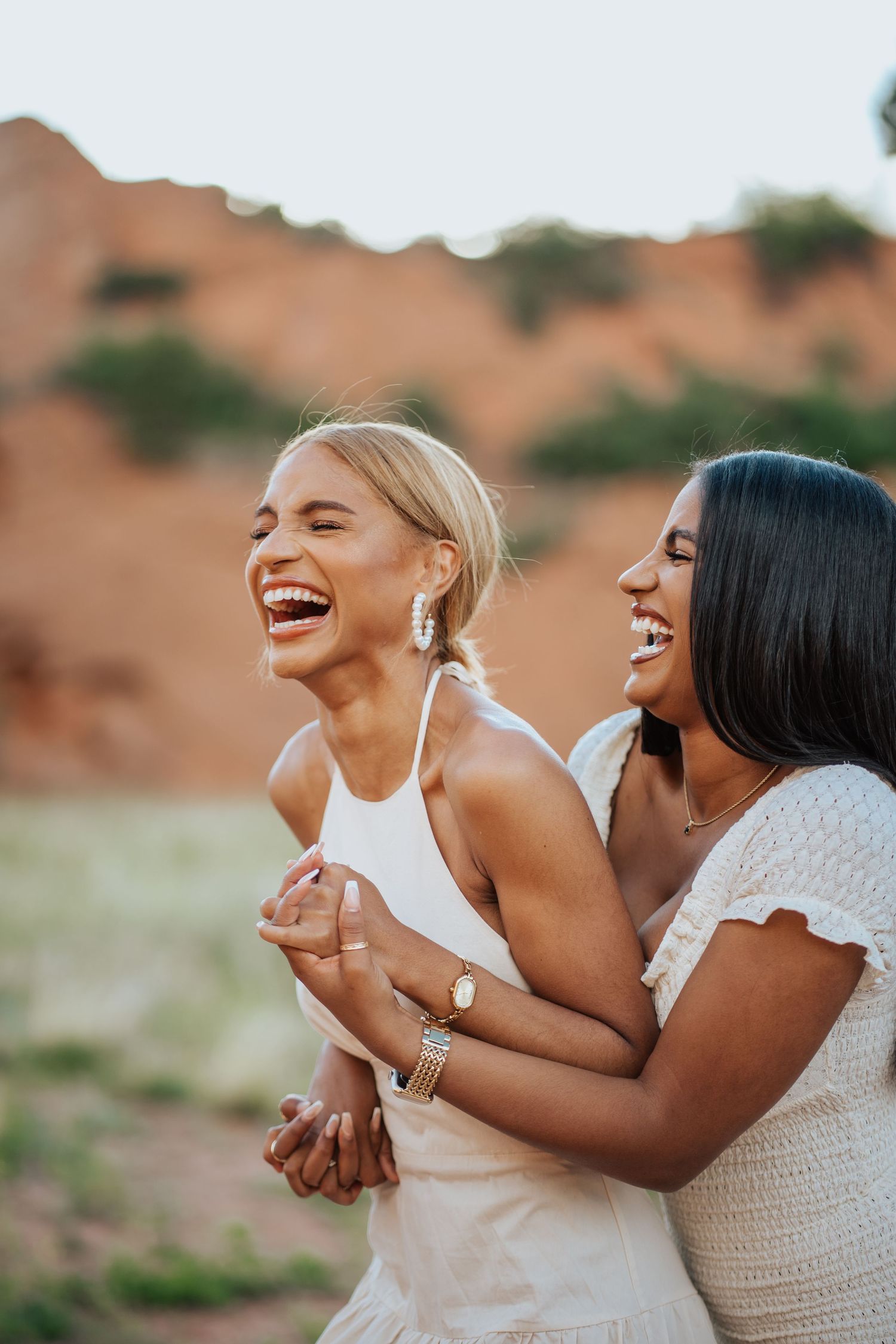Two women laughing joyfully outdoors near red rock formations in white summer dresses.