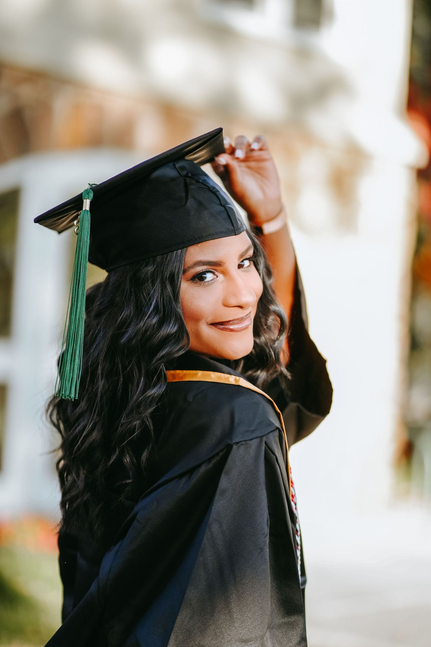 Young woman in black graduation cap and gown smiling over shoulder during outdoor graduation photo session.