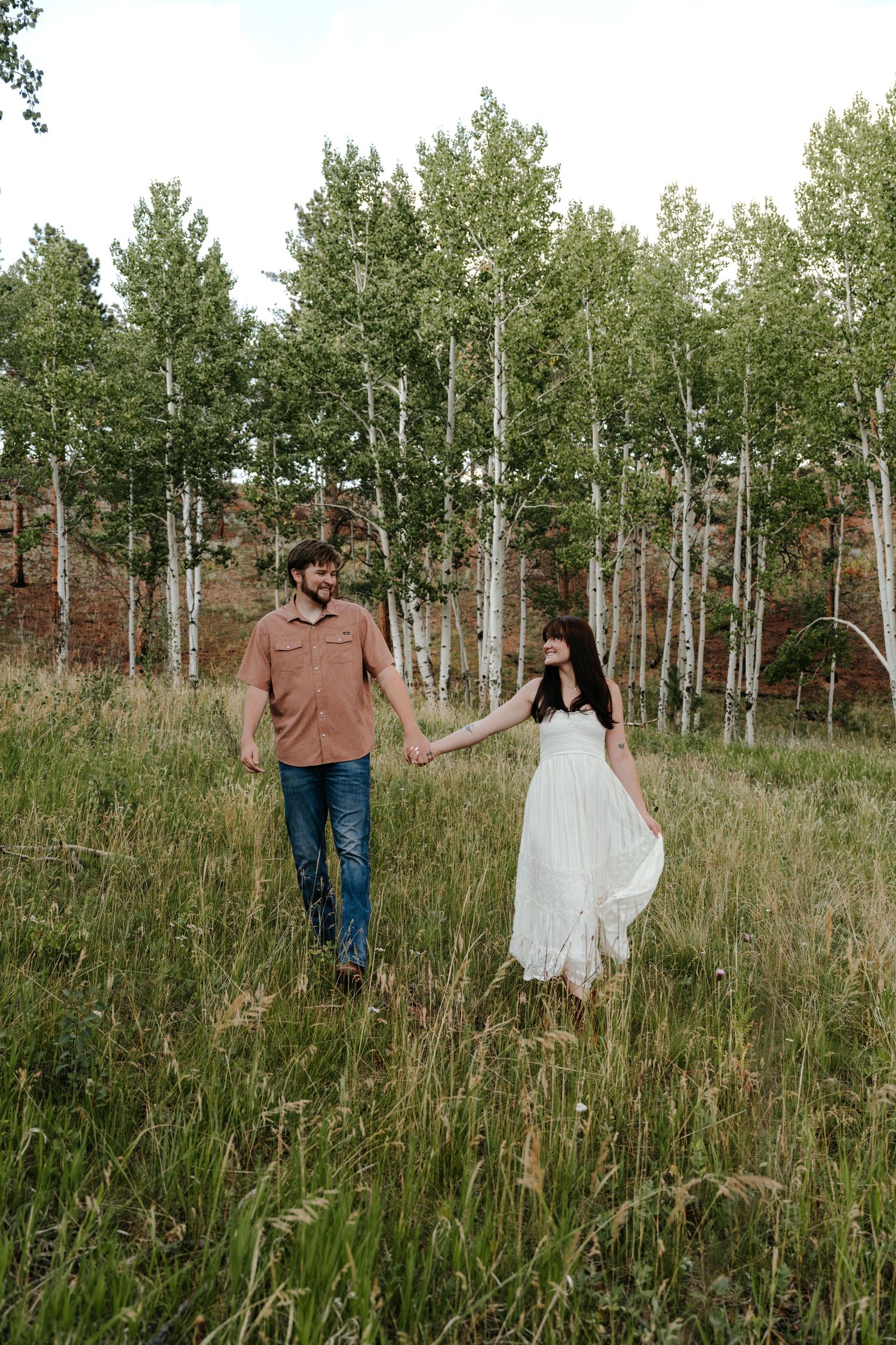 Couple holding hands walking through a green meadow with white birch trees in background.