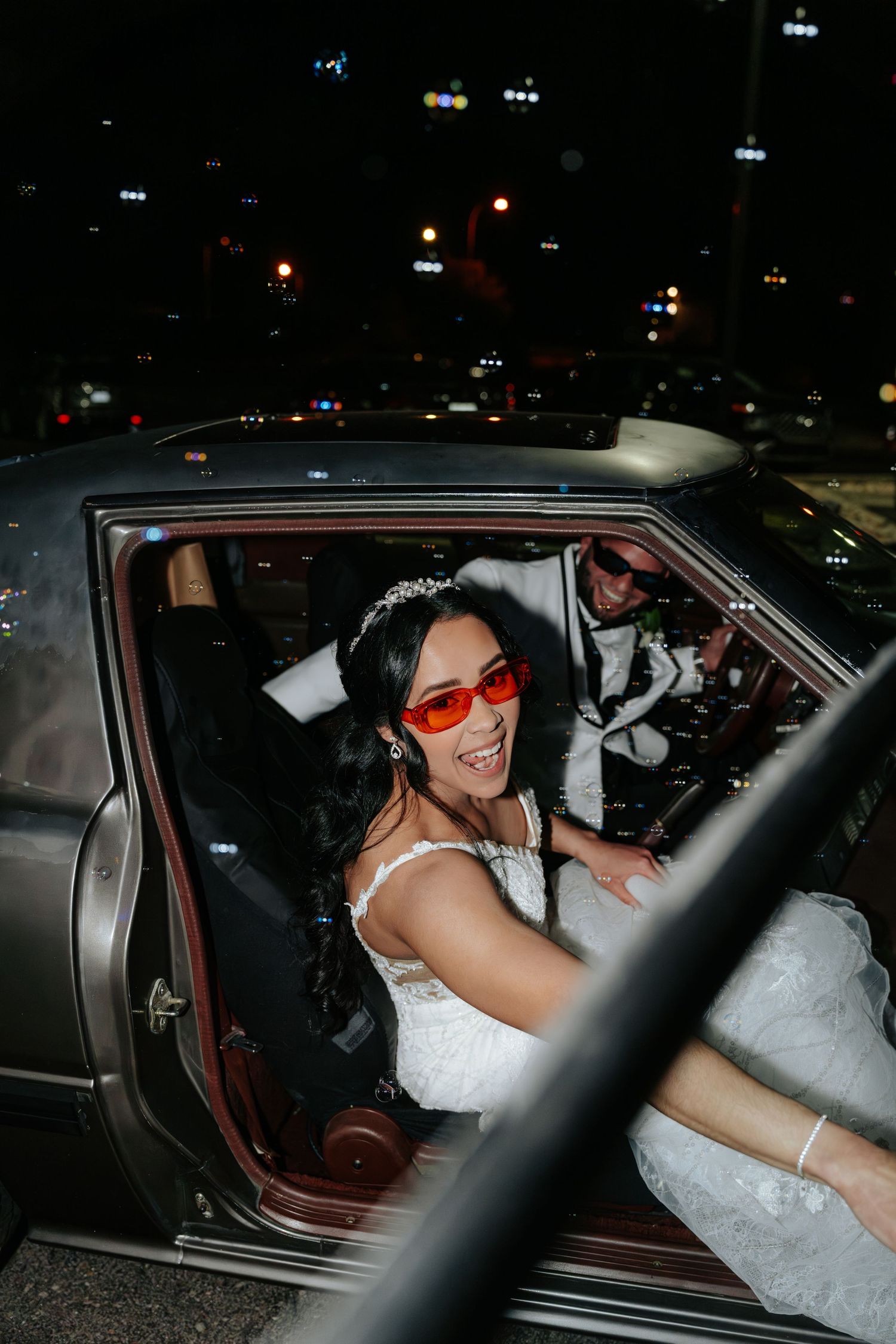 Joyful bride in wedding dress wearing red sunglasses laughs inside vintage car at night wedding celebration.