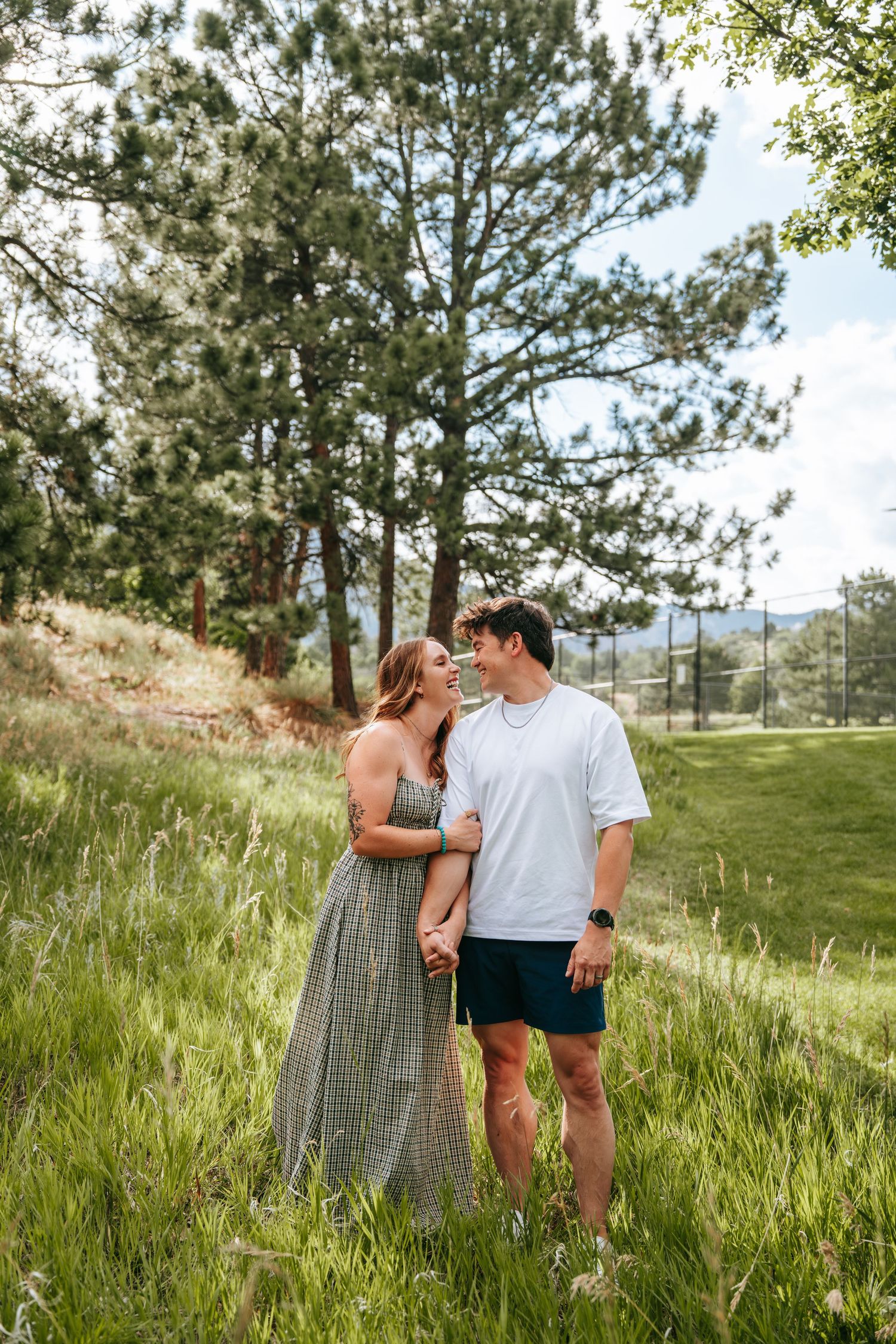 A couple smiles at each other in a lush green meadow surrounded by pine trees and mountains.