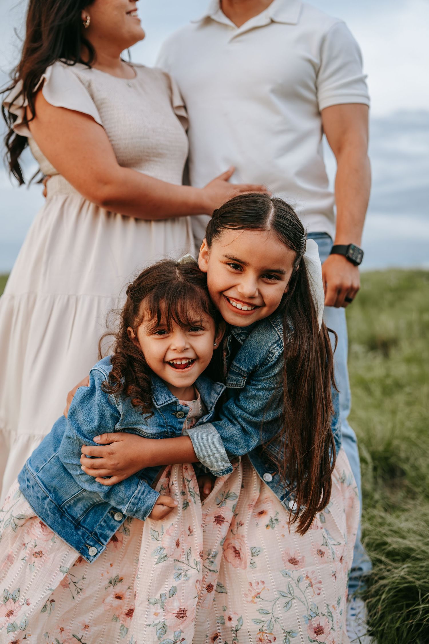 Two young sisters in denim jackets hug and smile with parents standing behind them outdoors.