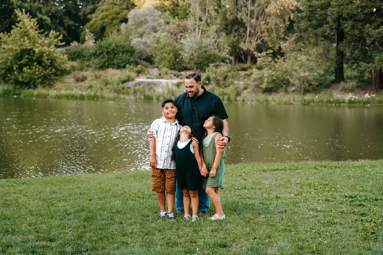 Father standing proudly with his three children on green grass in front of a calm pond.