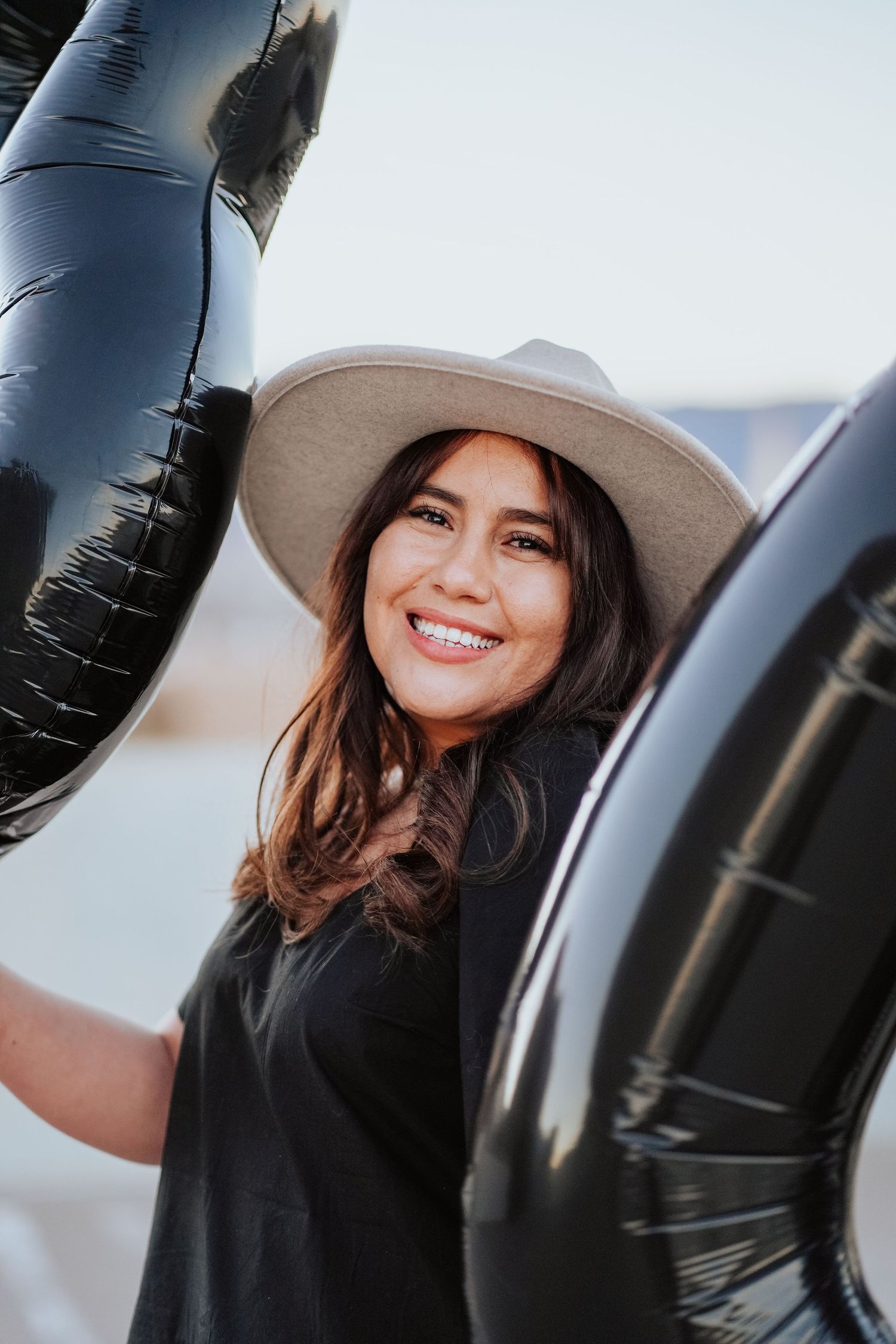 A smiling young woman in a white hat poses between large black number balloons outdoors.