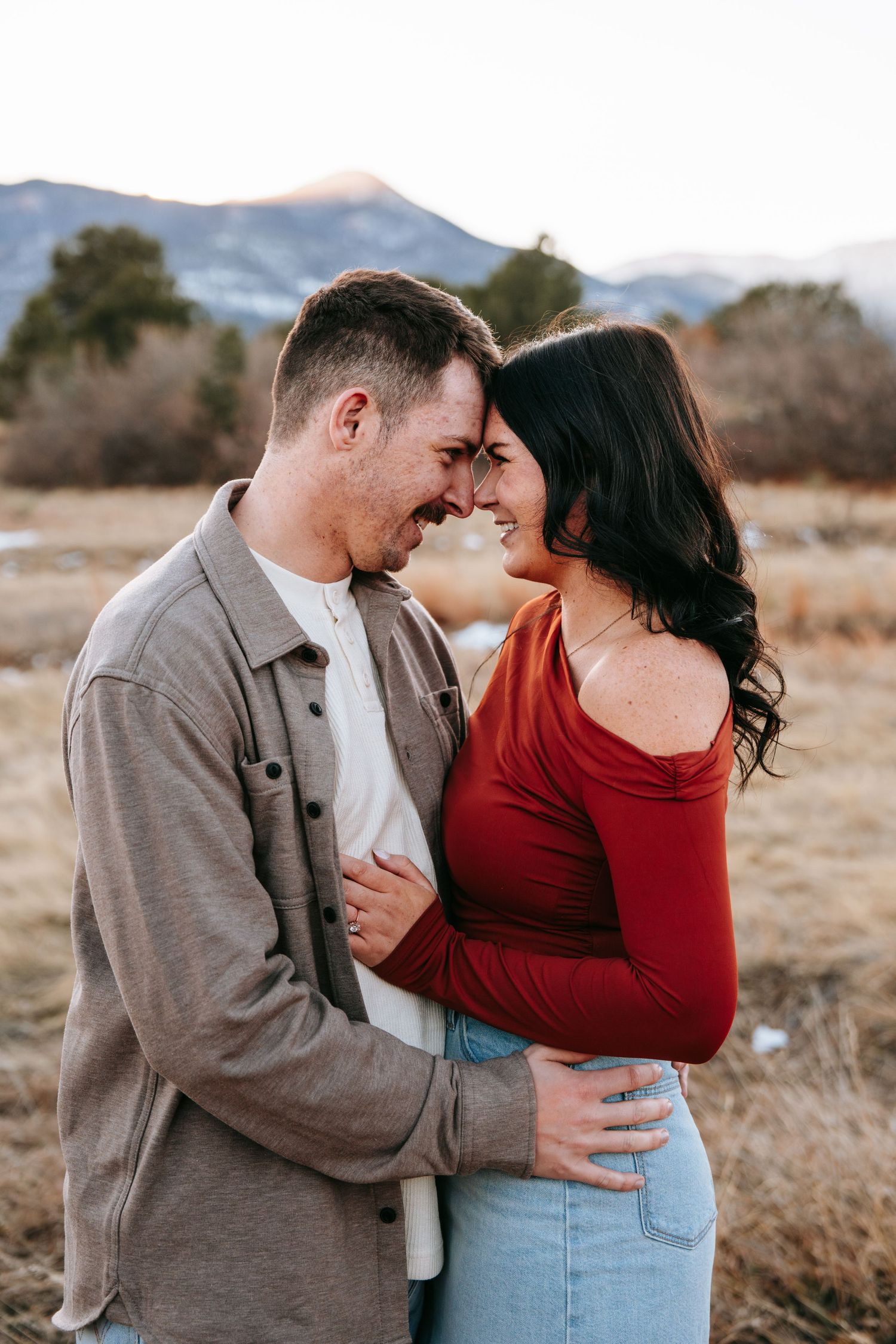 Couple laughing and embracing in a dry winter field with mountains in the background.