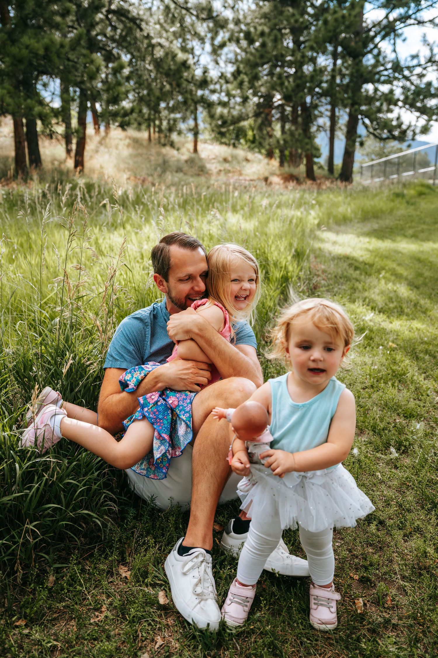 Father sitting on grass with two young blonde daughters, laughing together outdoors near pine trees.
