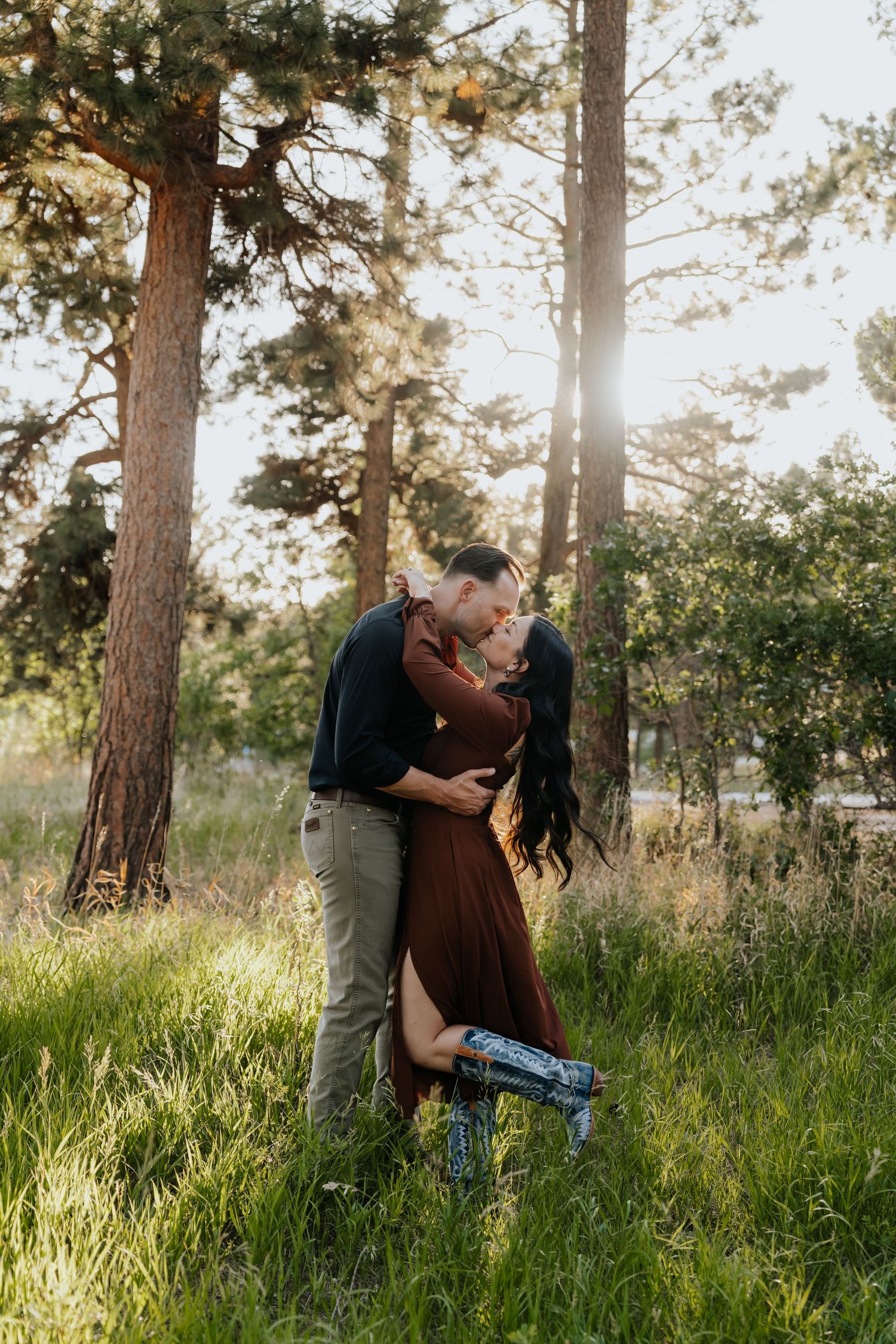 Couple sharing a romantic kiss in a sunlit forest clearing, woman in burgundy dress with one leg raised.