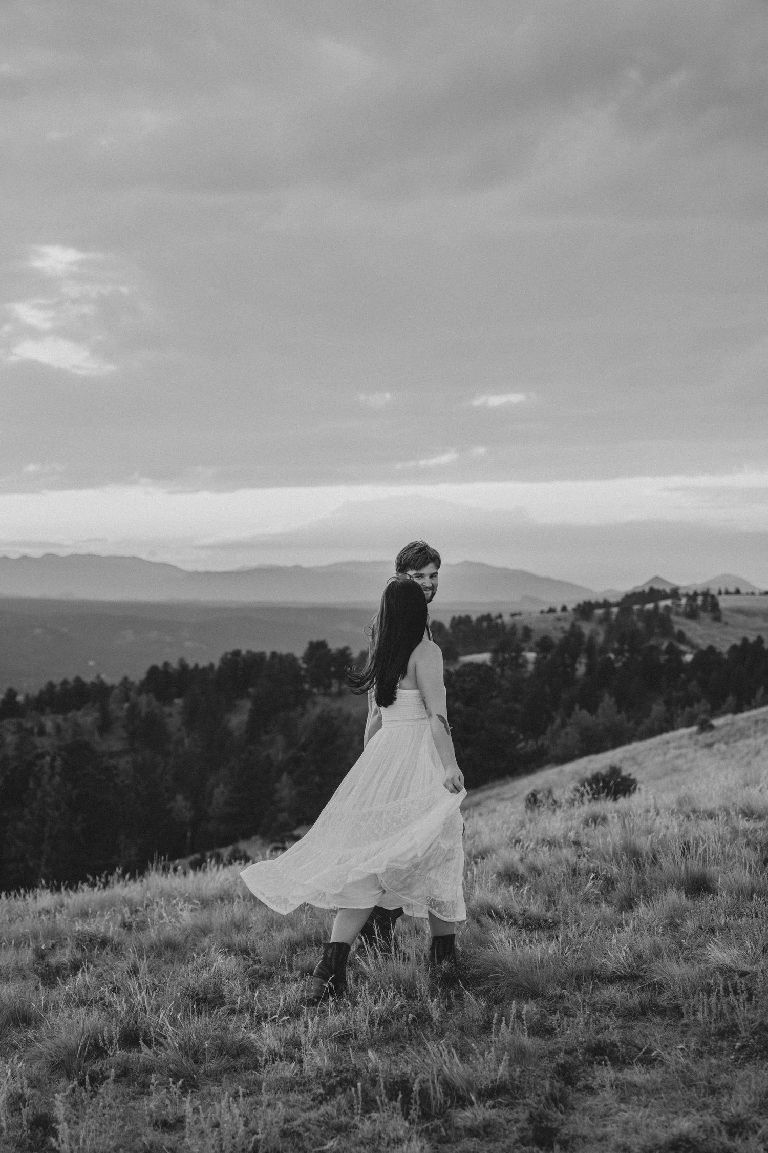 Black and white photo of a woman in a flowing white dress and boots standing on a hilltop with mountain views.