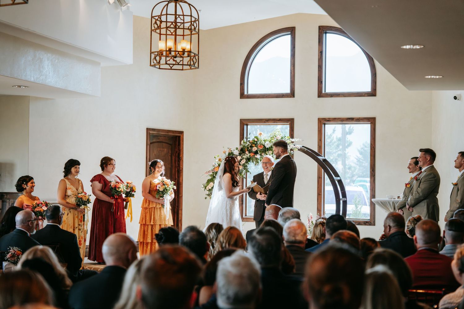 Wedding ceremony in elegant chapel with circular floral arch, bridesmaids in warm tones, and seated guests.
