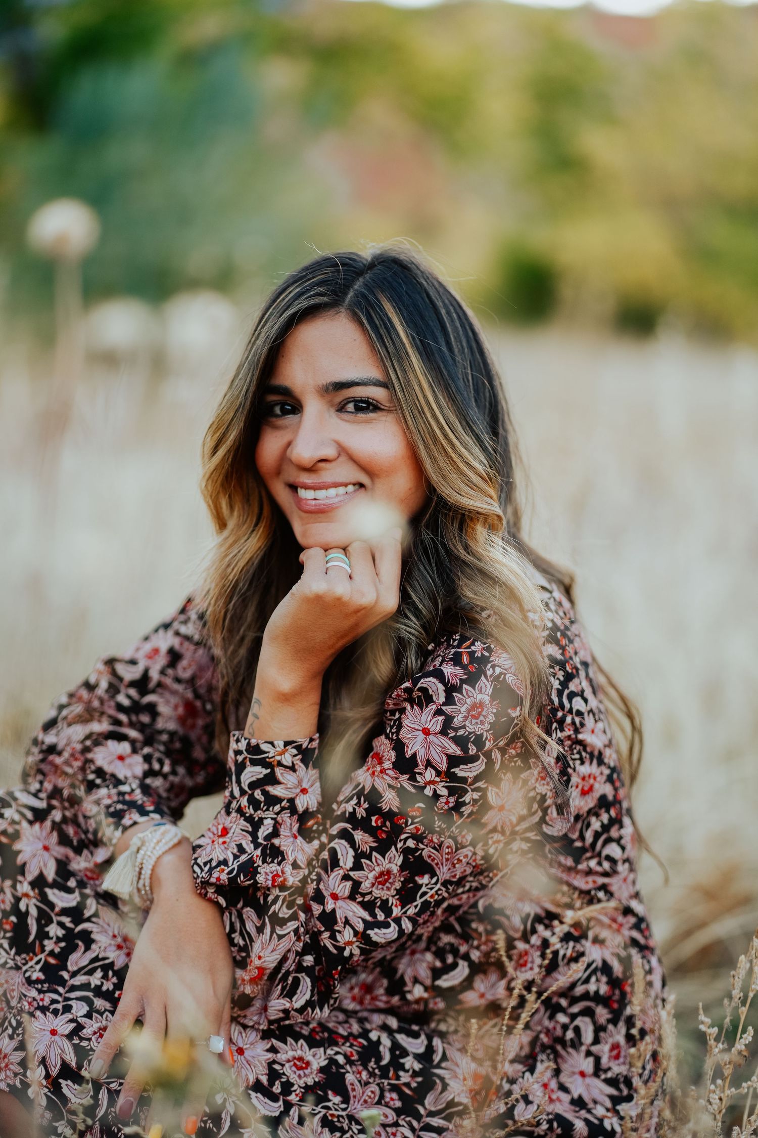 Cheerful brunette woman in floral dress resting chin on hand while smiling in golden field.