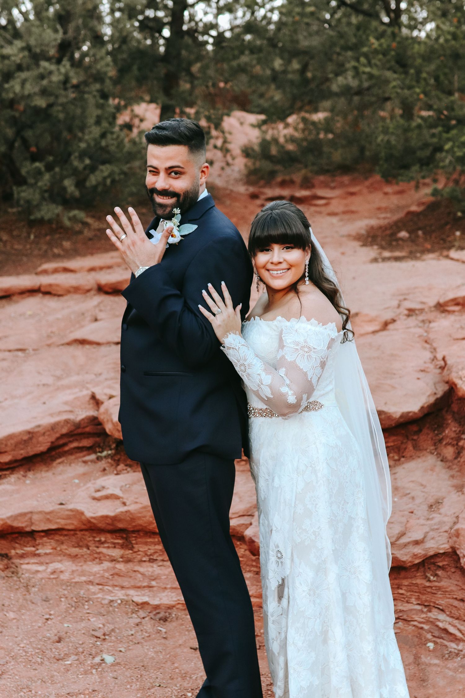 Bride and groom posing playfully on red rocks, groom waving at camera outdoors.