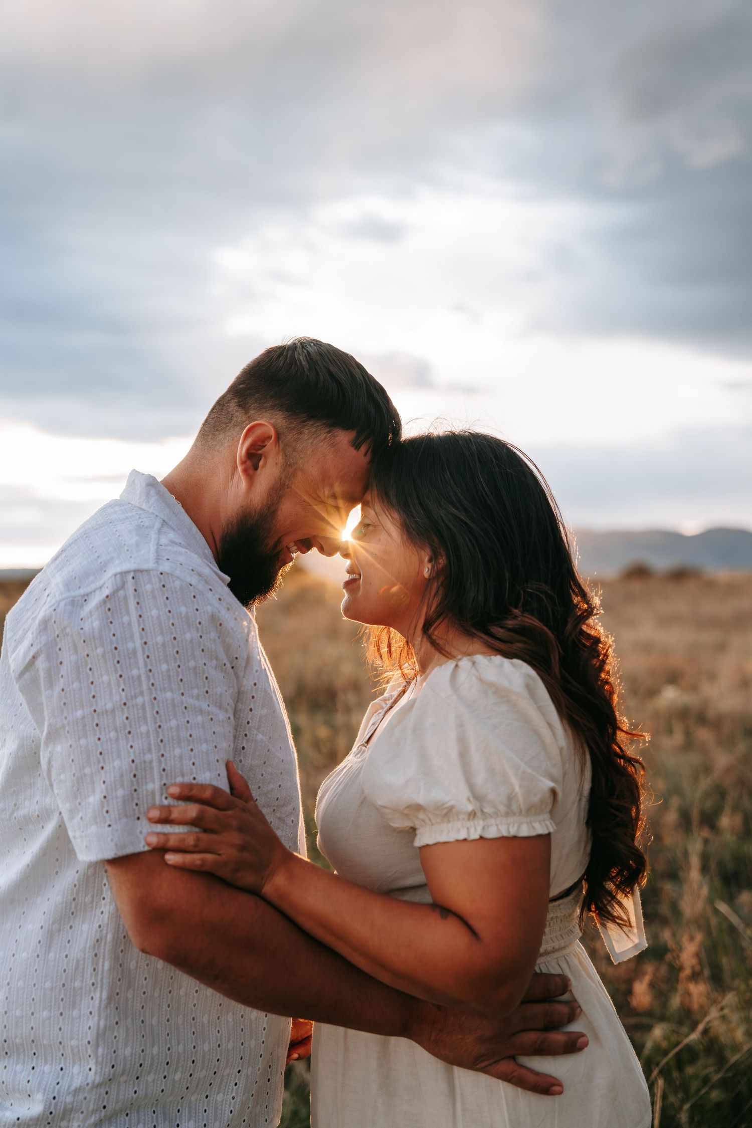 Couple in white outfits embrace forehead-to-forehead at golden sunset in open field.