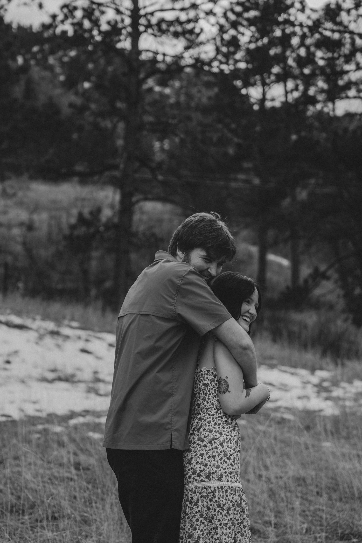 Black and white image of couple sharing a tender embrace in a pine forest meadow setting.