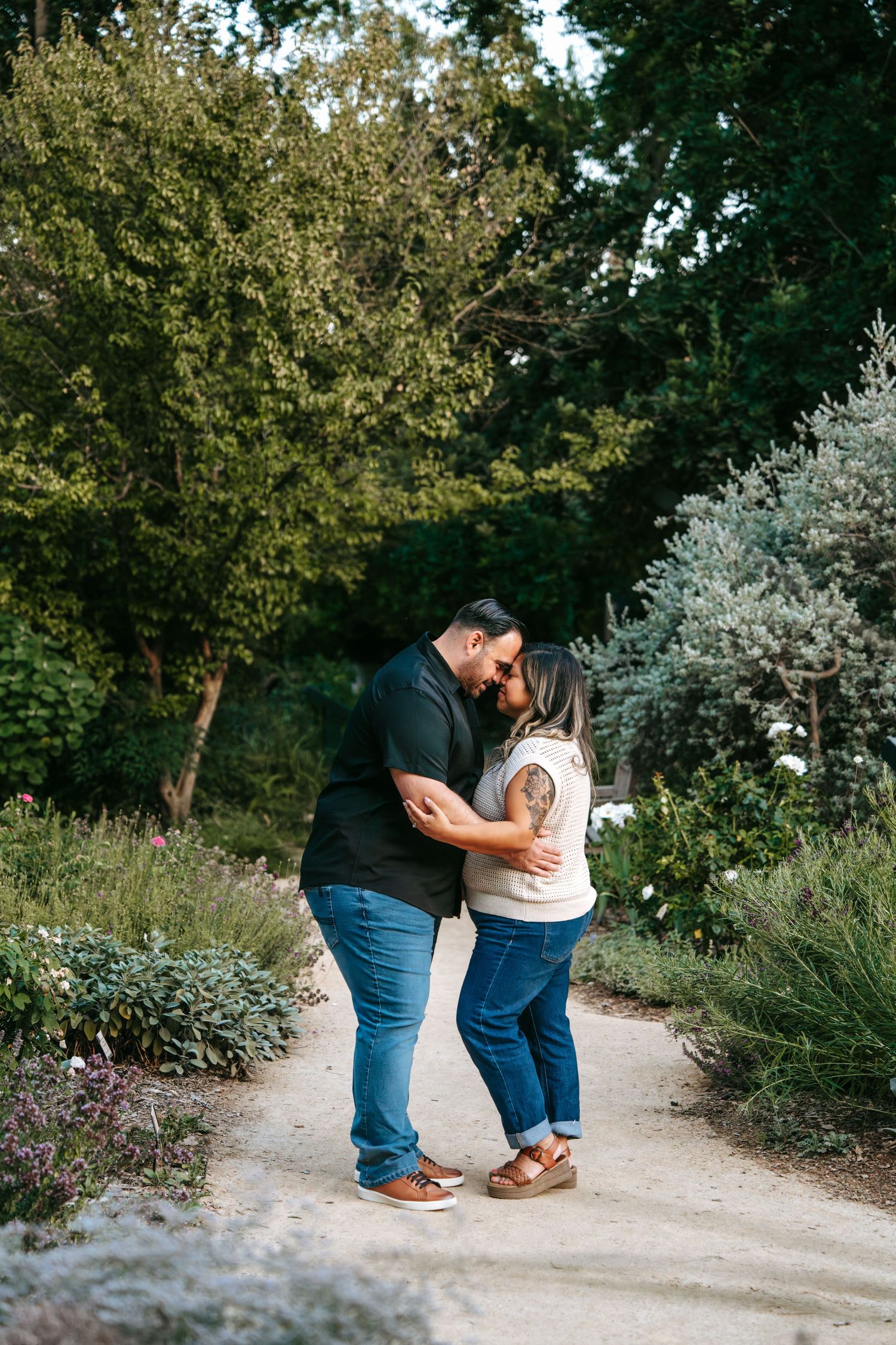 Romantic couple sharing a kiss on a garden path surrounded by lush green shrubs and trees.