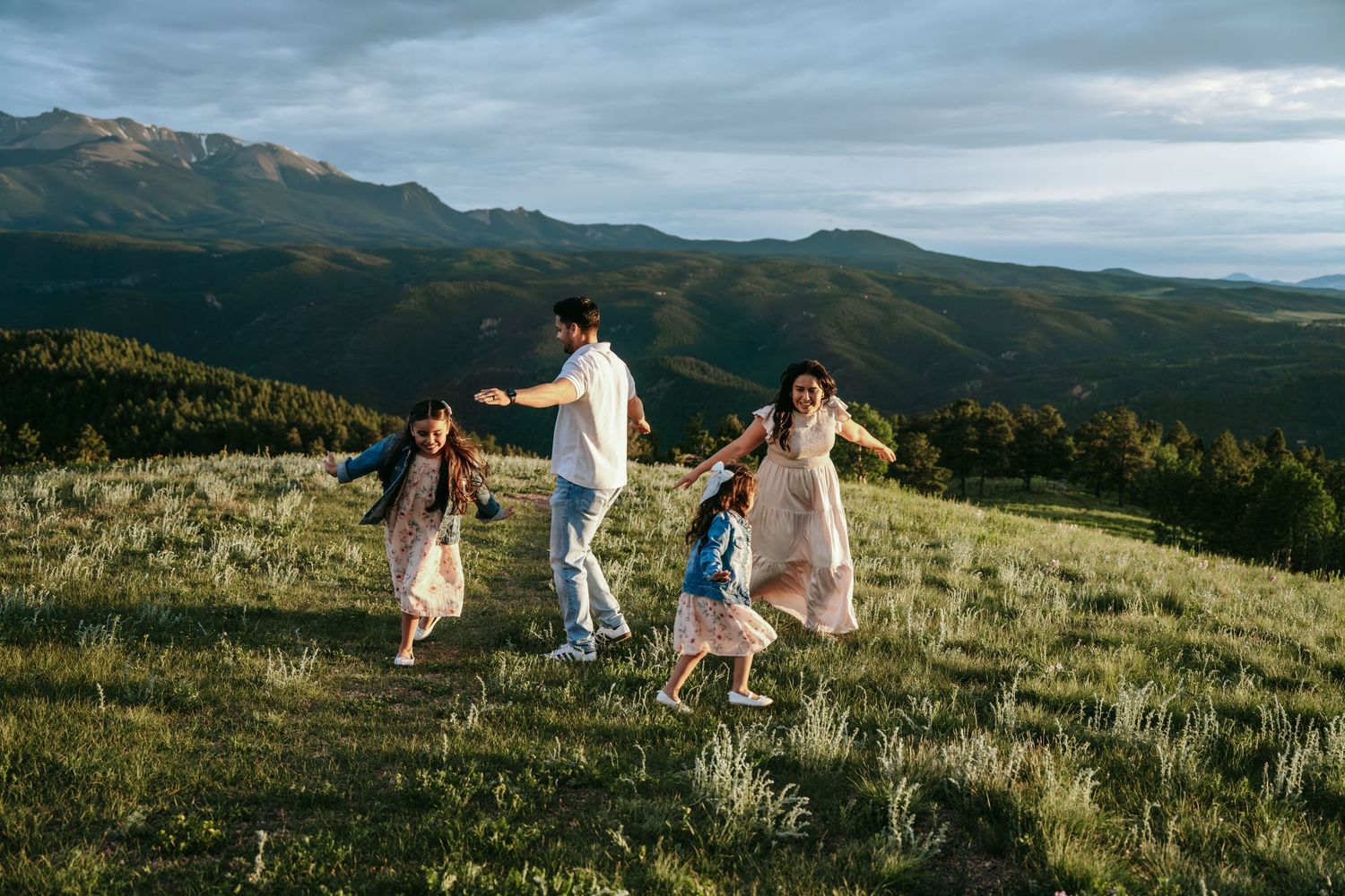 Family of four playing and running on a grassy mountain meadow at golden hour with scenic mountain views.