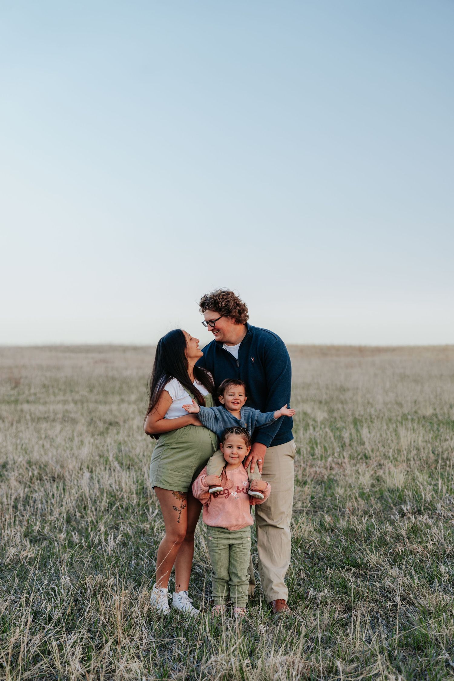 Happy family of four posing together in an open grassy field at dusk.