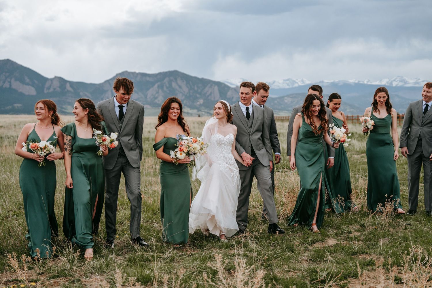 Wedding party in gray suits and emerald green dresses walking through mountain meadow under cloudy skies.