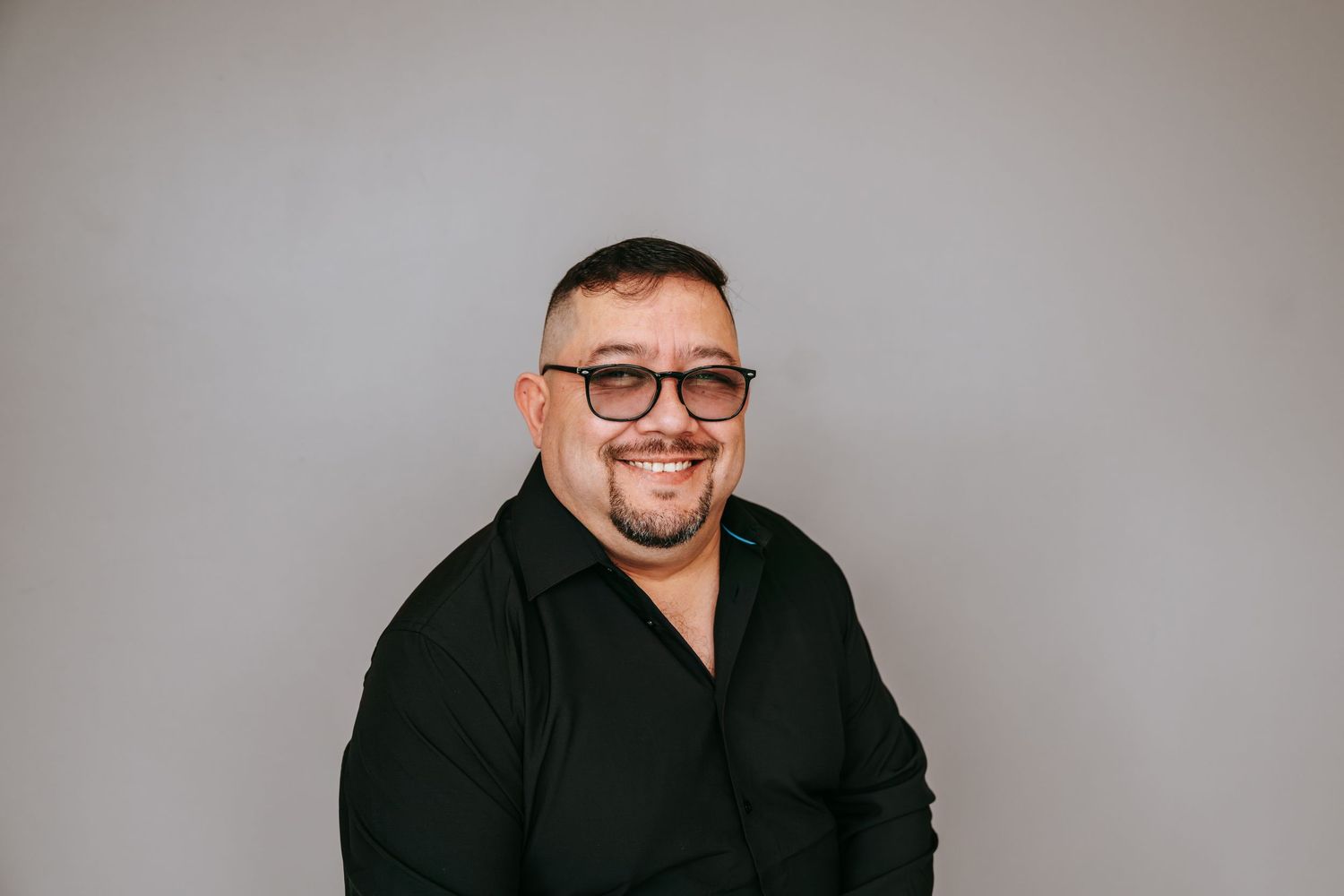 Smiling man with glasses wearing a black shirt seated against a light gray background.