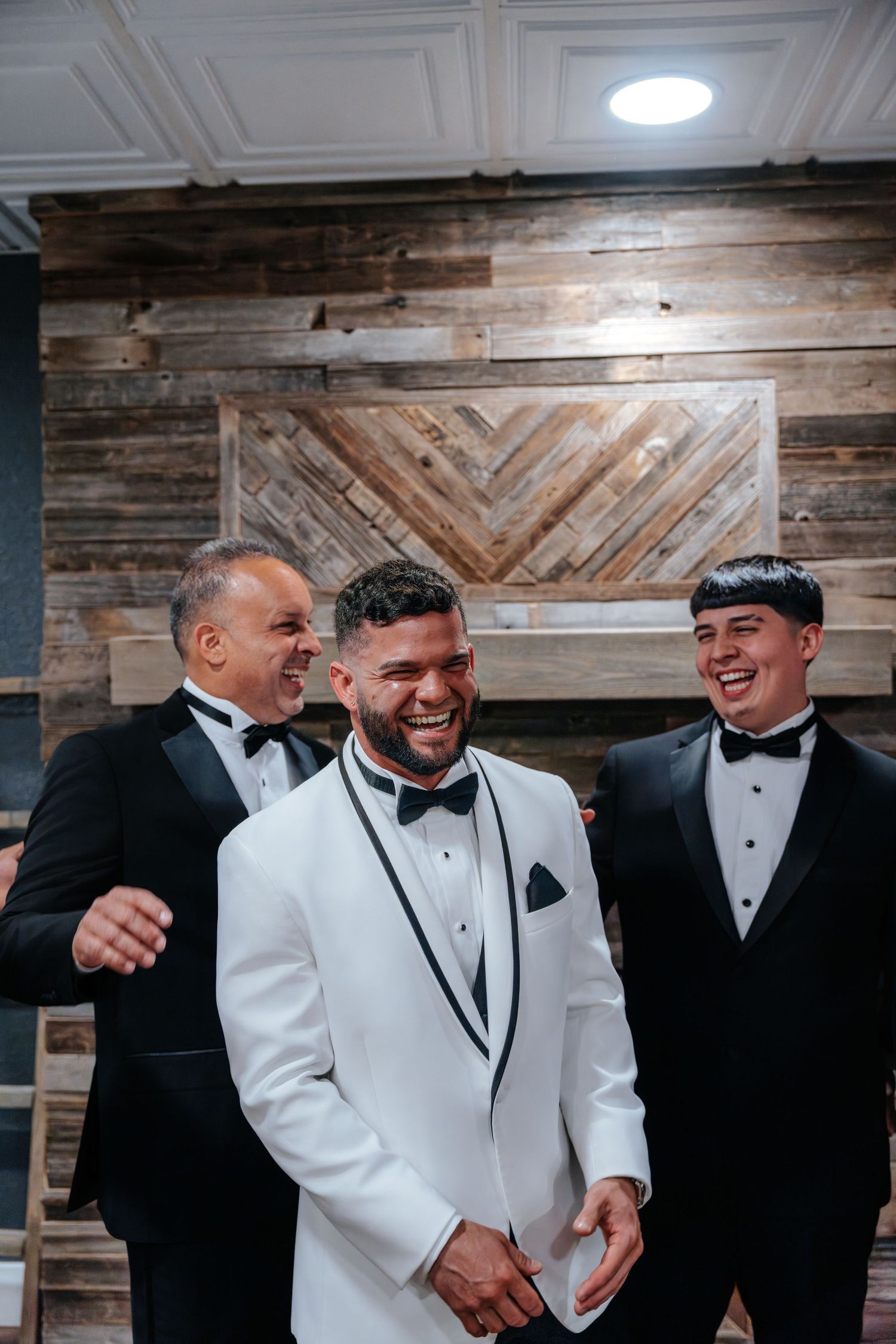Groom in white tuxedo laughing with two groomsmen in black tuxedos before wedding ceremony.