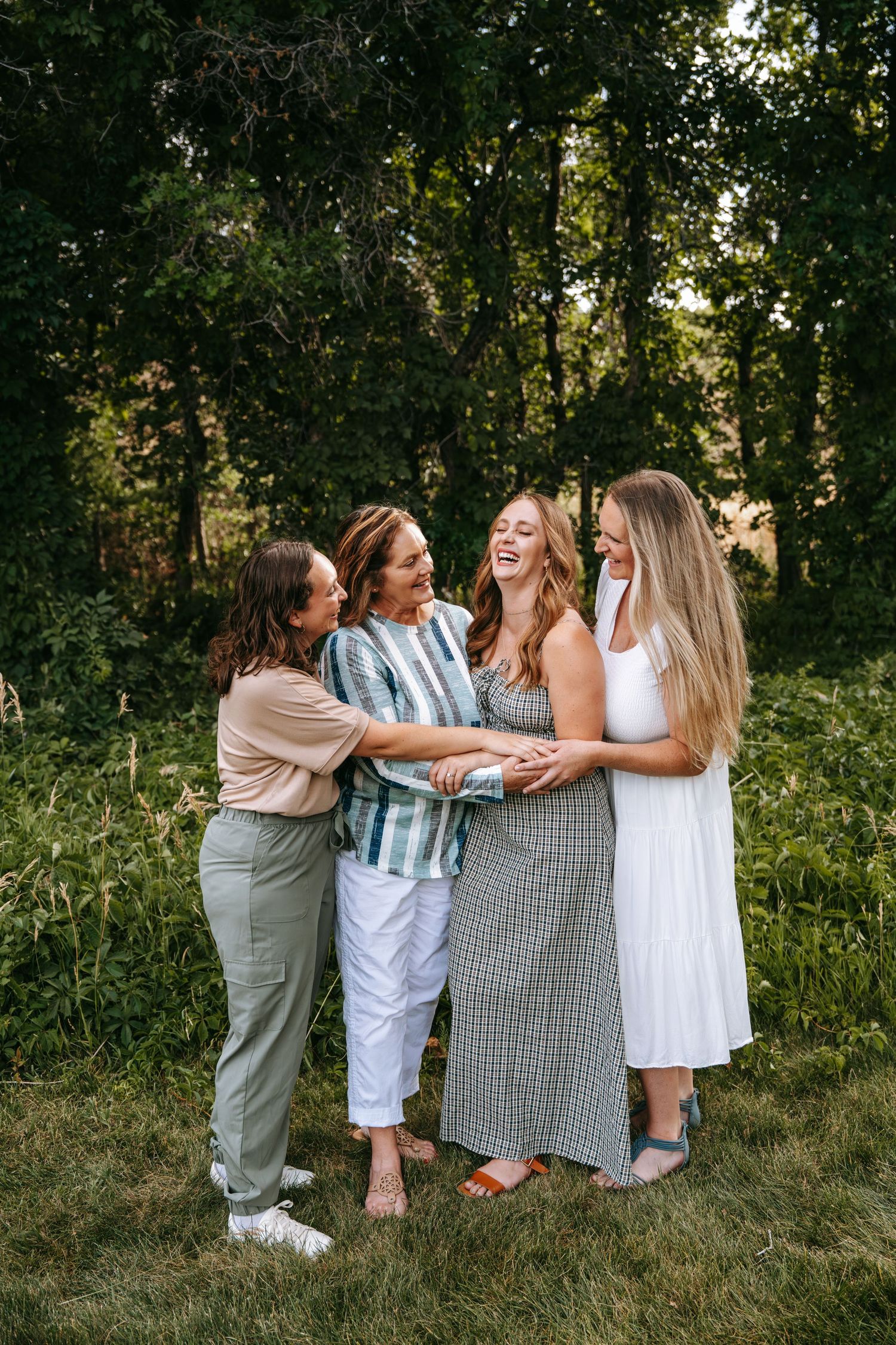 Four women laughing and embracing outdoors in a lush green setting, wearing casual summer outfits.