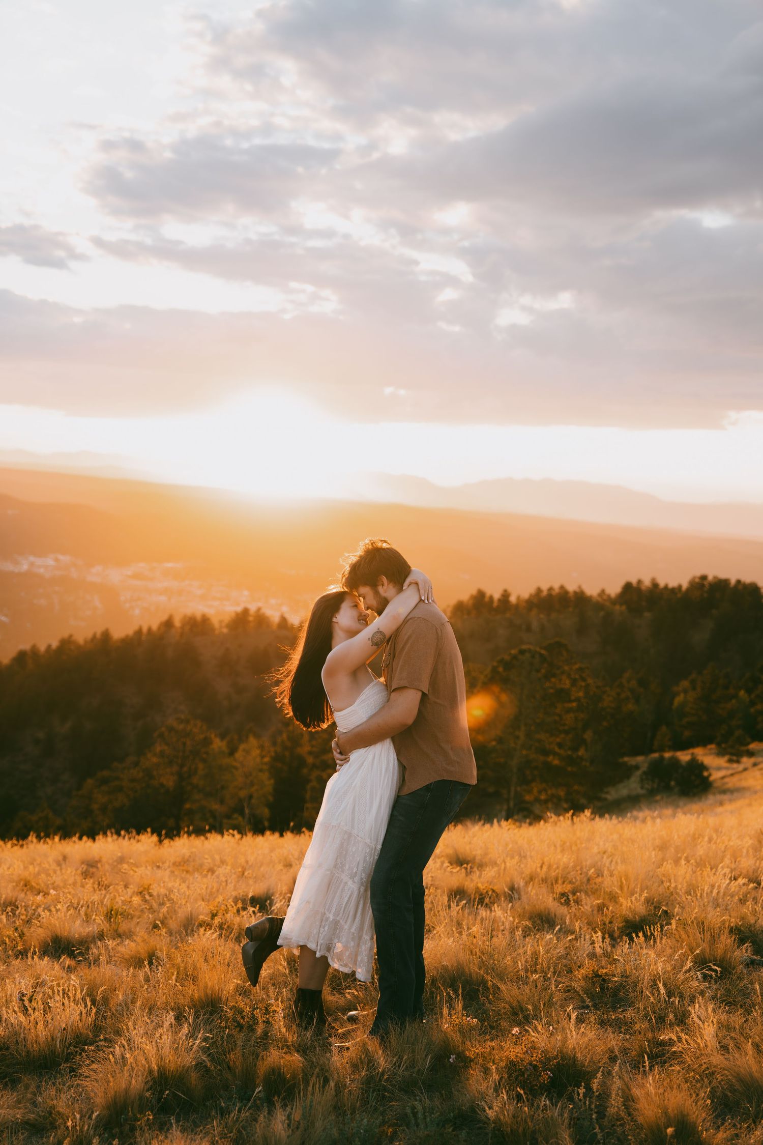 Couple embracing at golden sunset on a hilltop meadow with mountain forest views in warm evening light.
