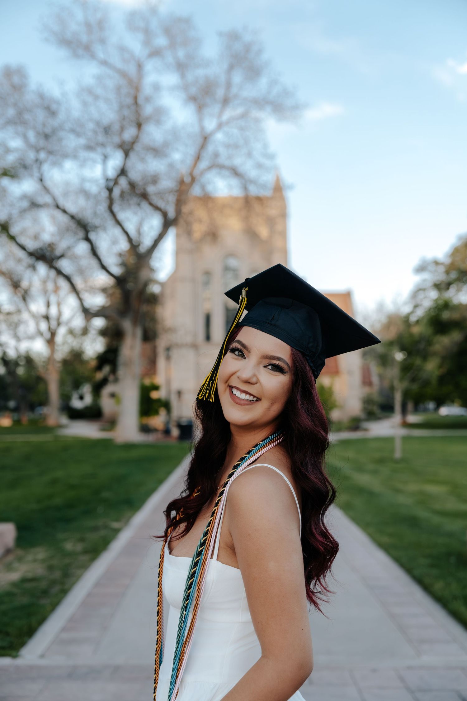 Happy female graduate in cap and white dress smiles on a sunny college campus.