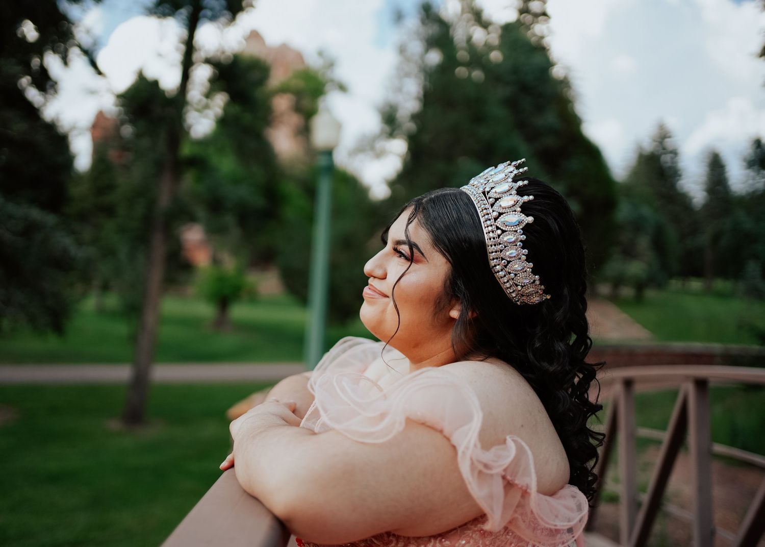 A young woman in a pink quinceañera gown and sparkling tiara gazes upward in a lush park.