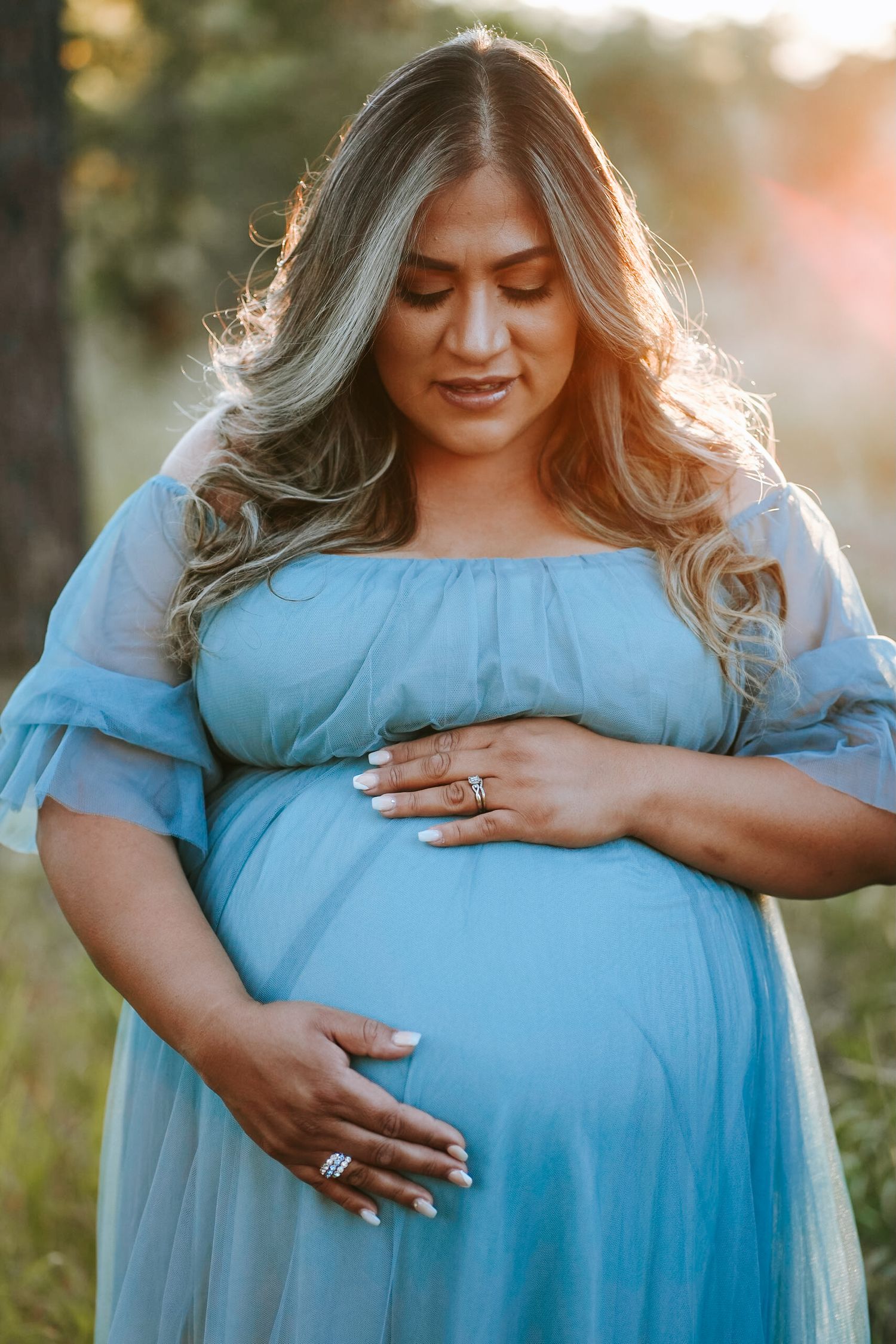 Close-up of pregnant woman in blue dress gently holding her baby bump outdoors.