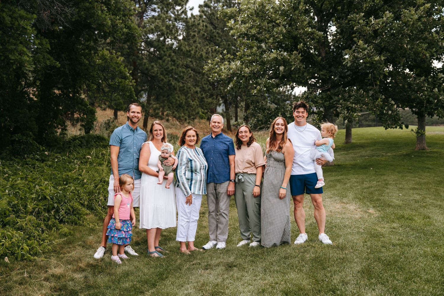 Large family group portrait smiling outdoors on green lawn surrounded by trees on a sunny day.