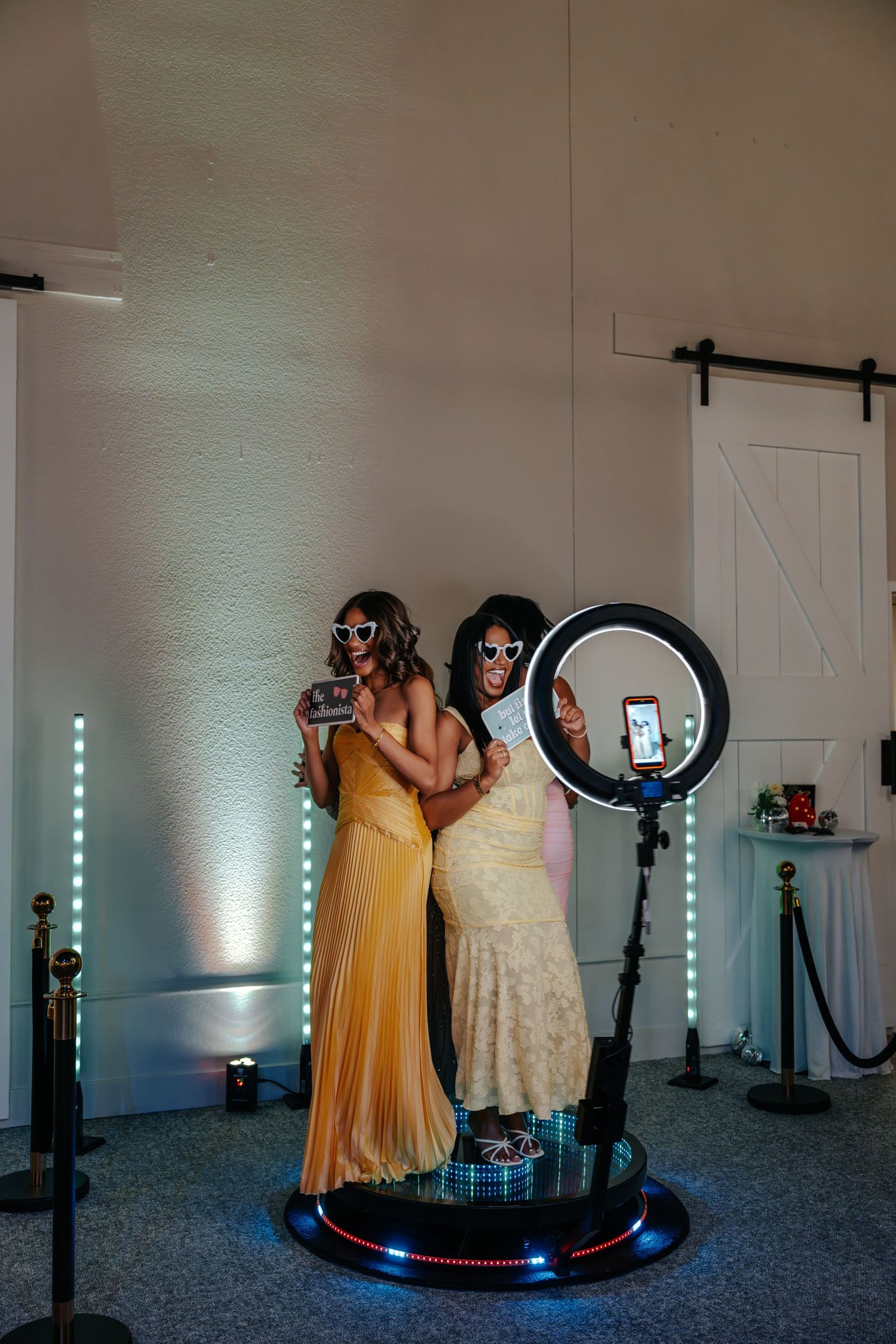 Two women in elegant gowns pose on a 360 photo booth platform with a ring light at an event venue.