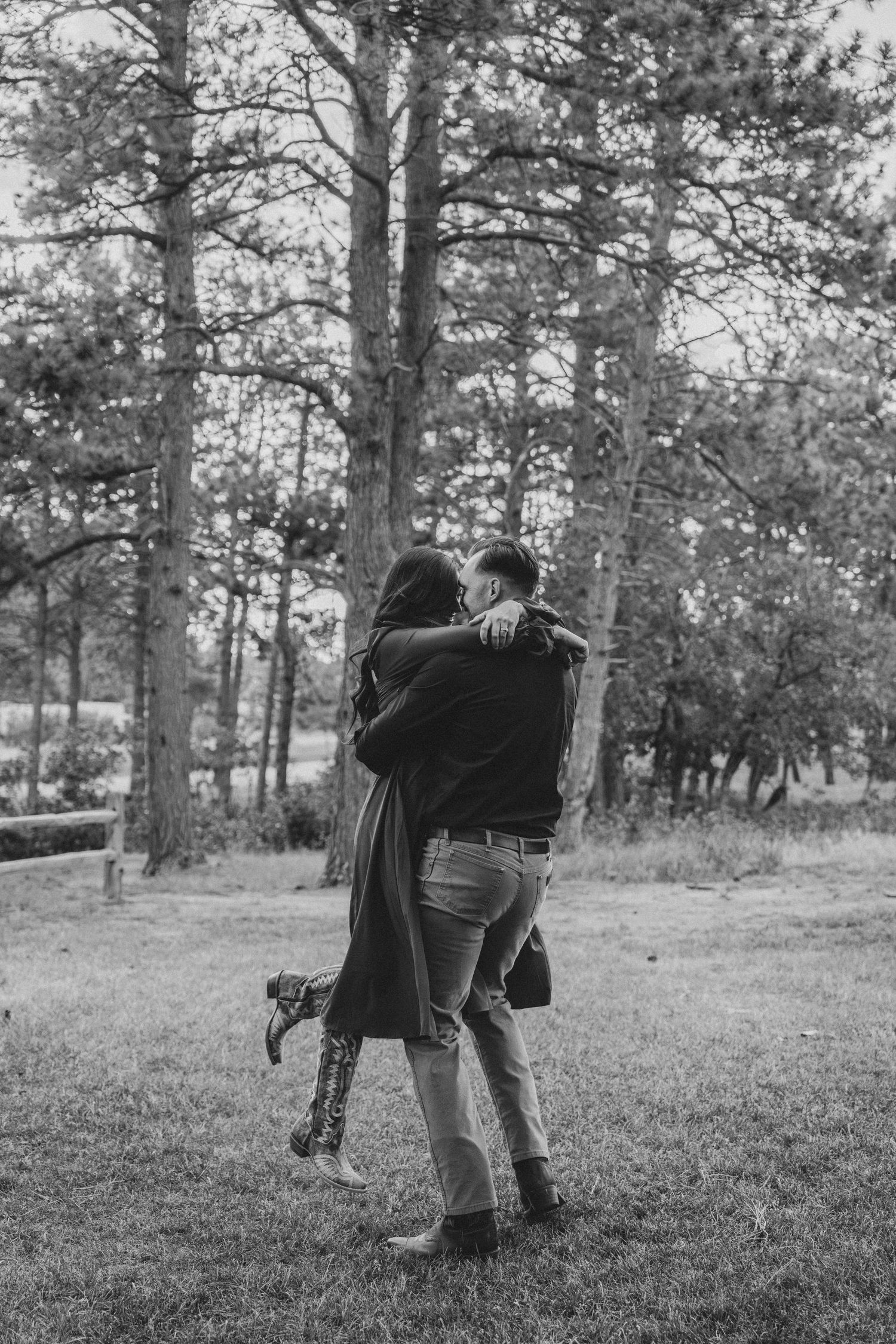 Black and white photo of a couple embracing and kissing outdoors among tall trees on a grassy lawn.