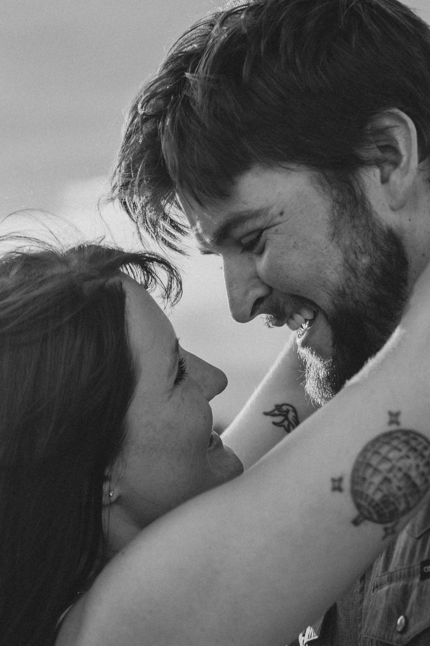 Black and white photo of a smiling couple gazing at each other, man with beard and tattoo embracing woman outdoors.
