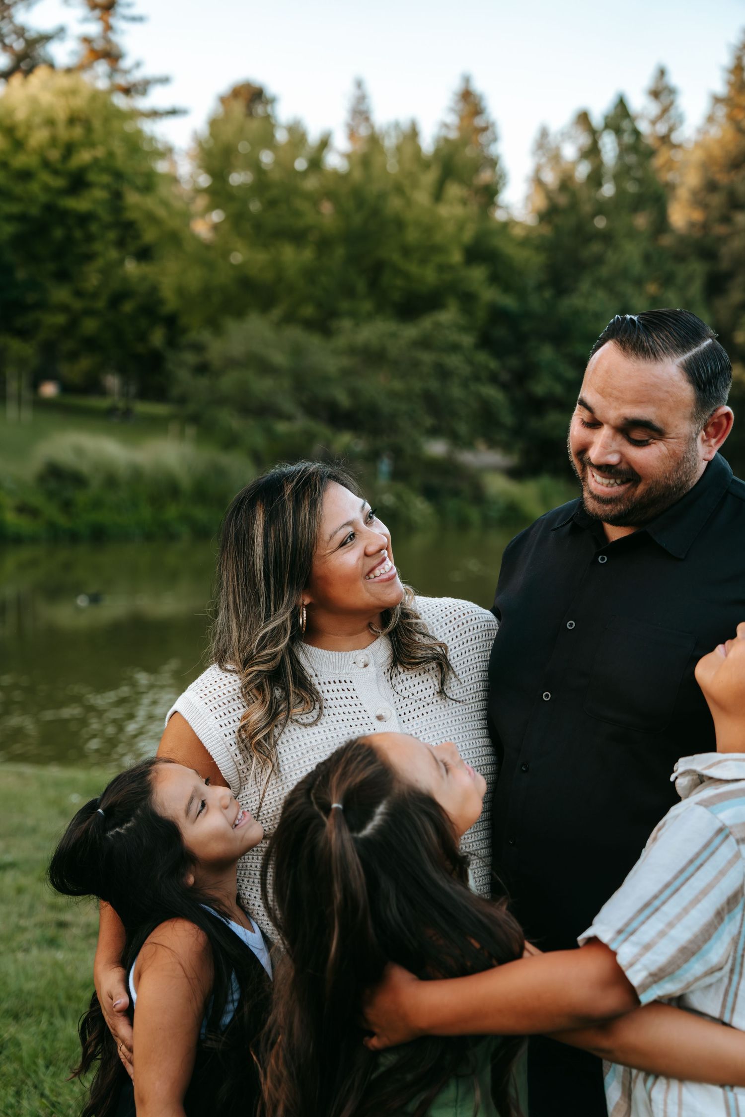 Joyful couple laughing while their children playfully hang on them near a peaceful park pond.