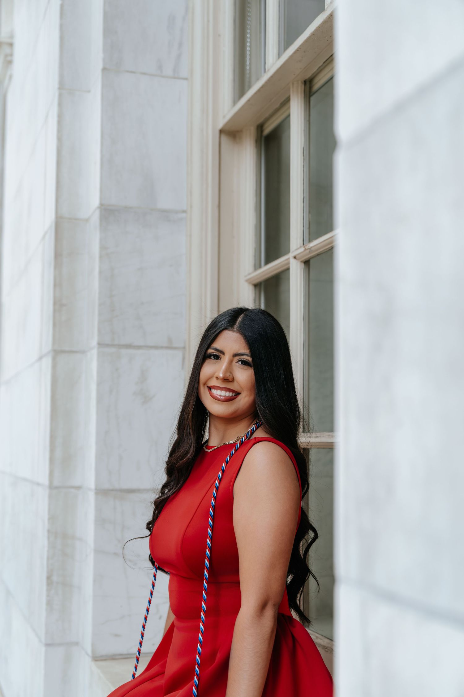 Smiling woman in red dress with graduation cords standing between marble columns by window.