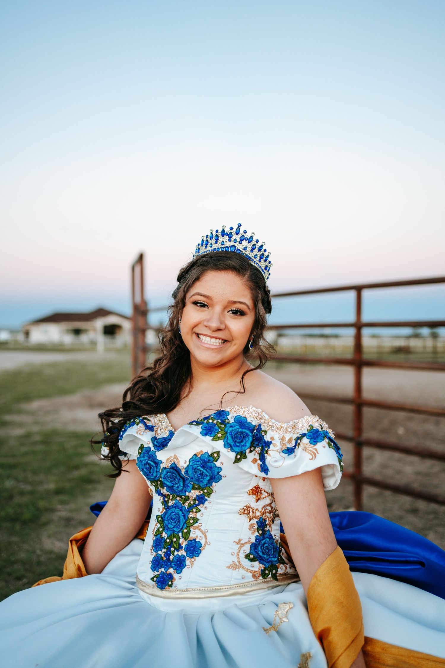 Joyful quinceañera in blue floral ball gown and tiara smiles at ranch fence at sunset.