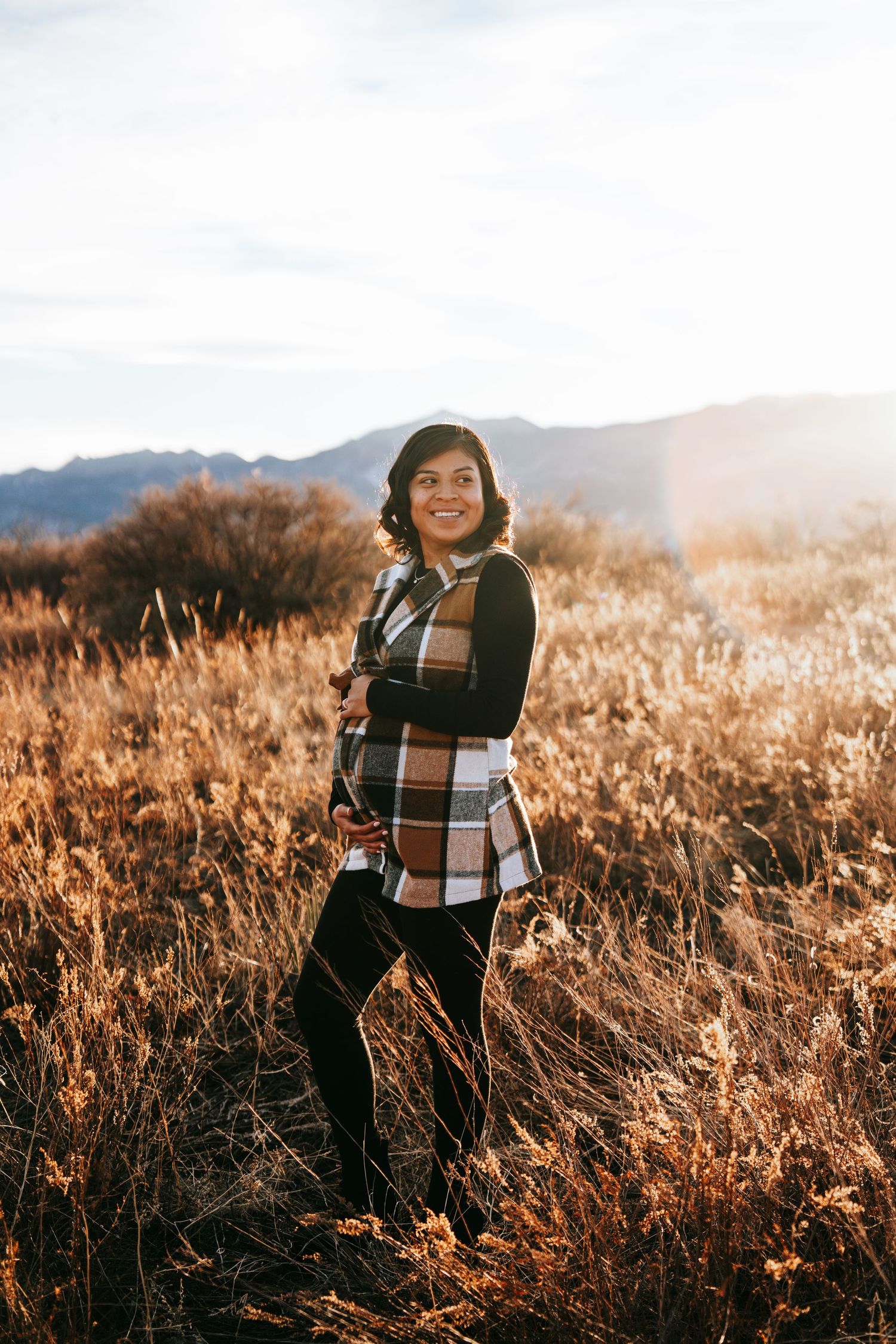 Smiling pregnant woman in plaid vest standing in a sunlit autumn field with mountains in background.