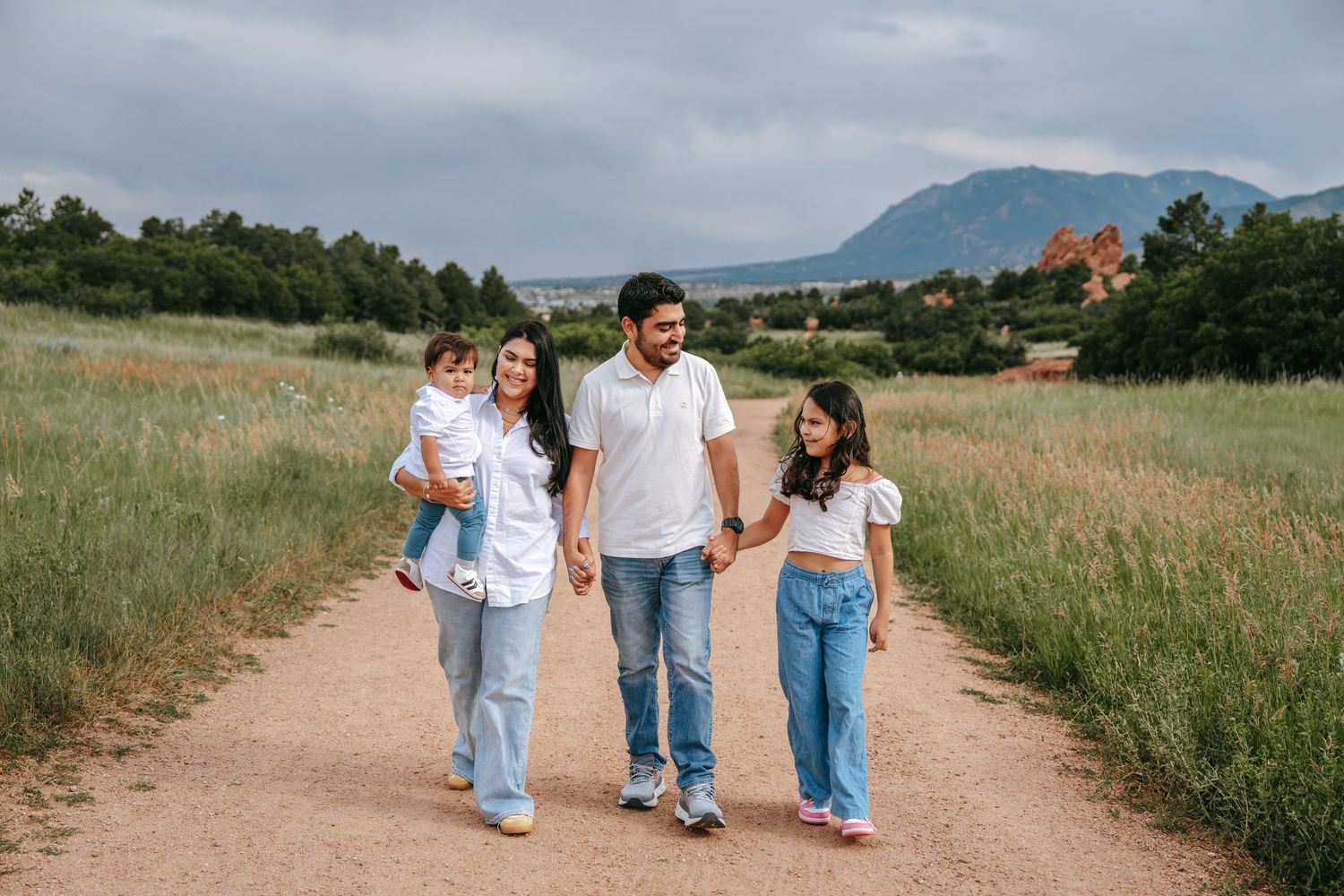 Happy family of four walking hand in hand along a scenic dirt path with mountains and red rocks behind them.