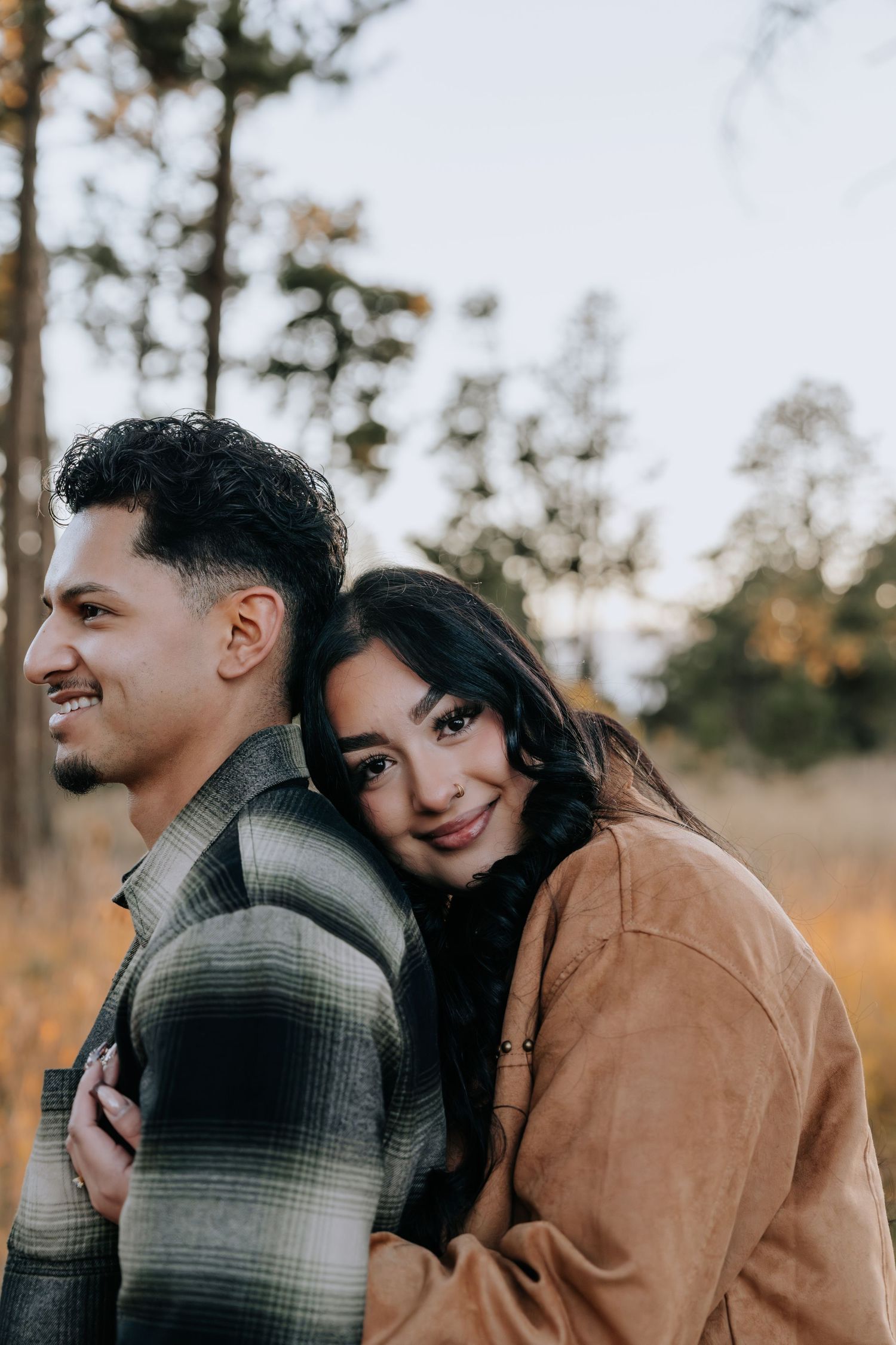 Woman rests her head on her partner's shoulder smiling in a golden autumn forest setting.