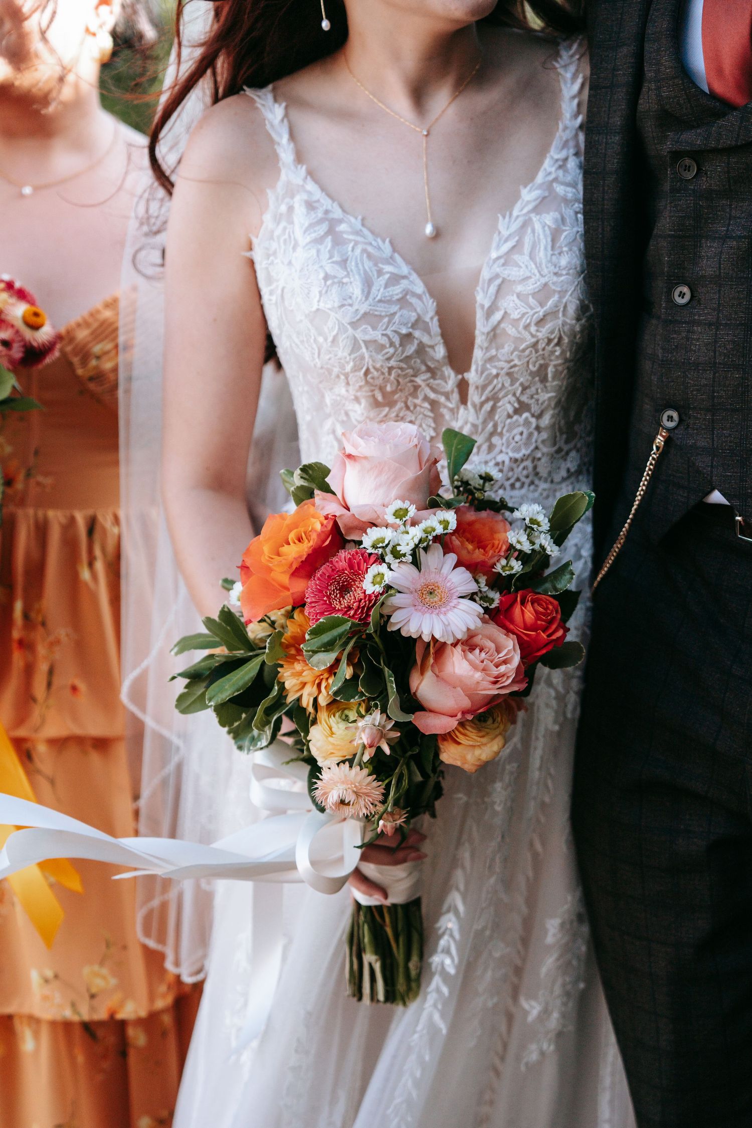 Close-up of bride in lace gown holding a vibrant orange, pink, and green wedding bouquet with ribbons.
