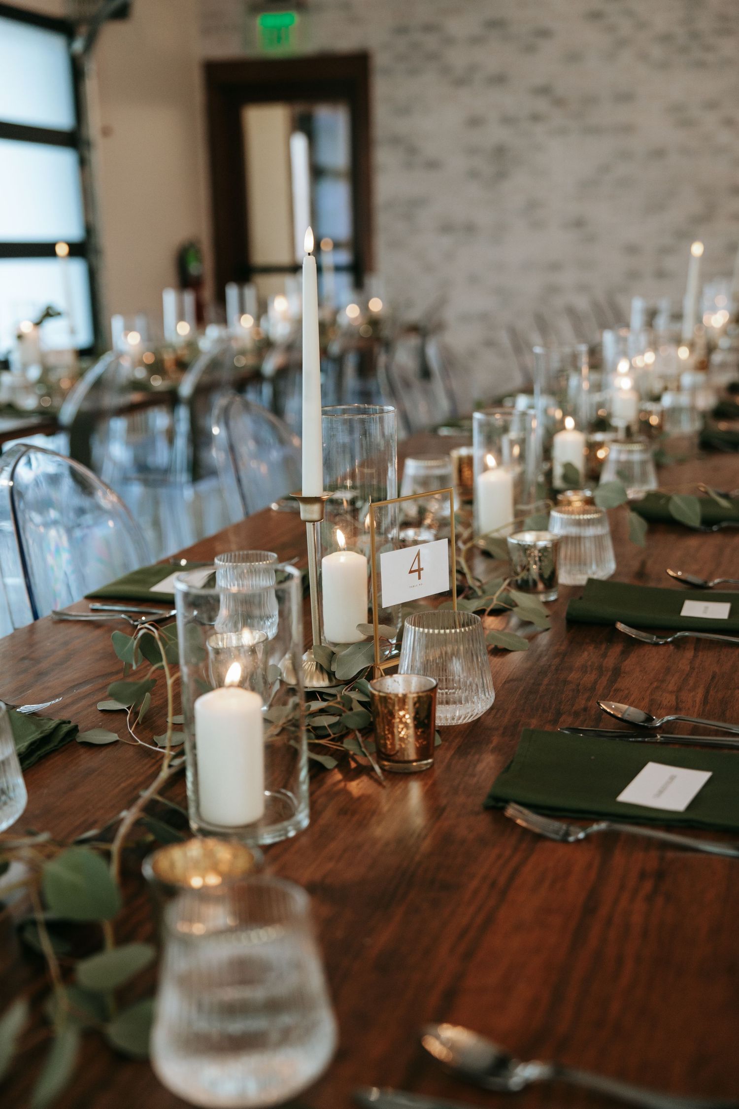 Long wooden reception table with white candles, eucalyptus garland, and green linen napkins.