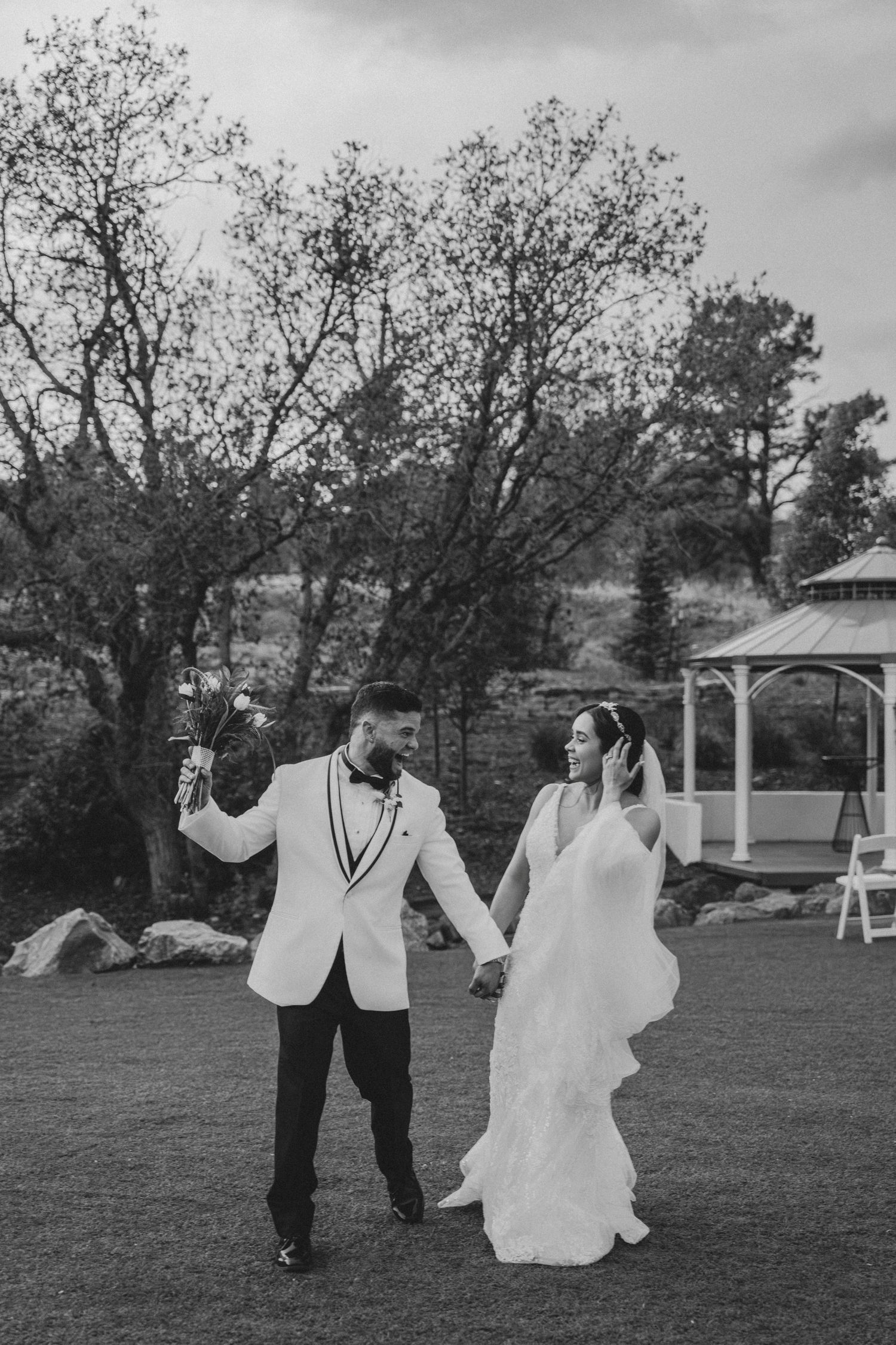 Black and white photo of a bride and groom holding hands walking on grass with a gazebo in the background.