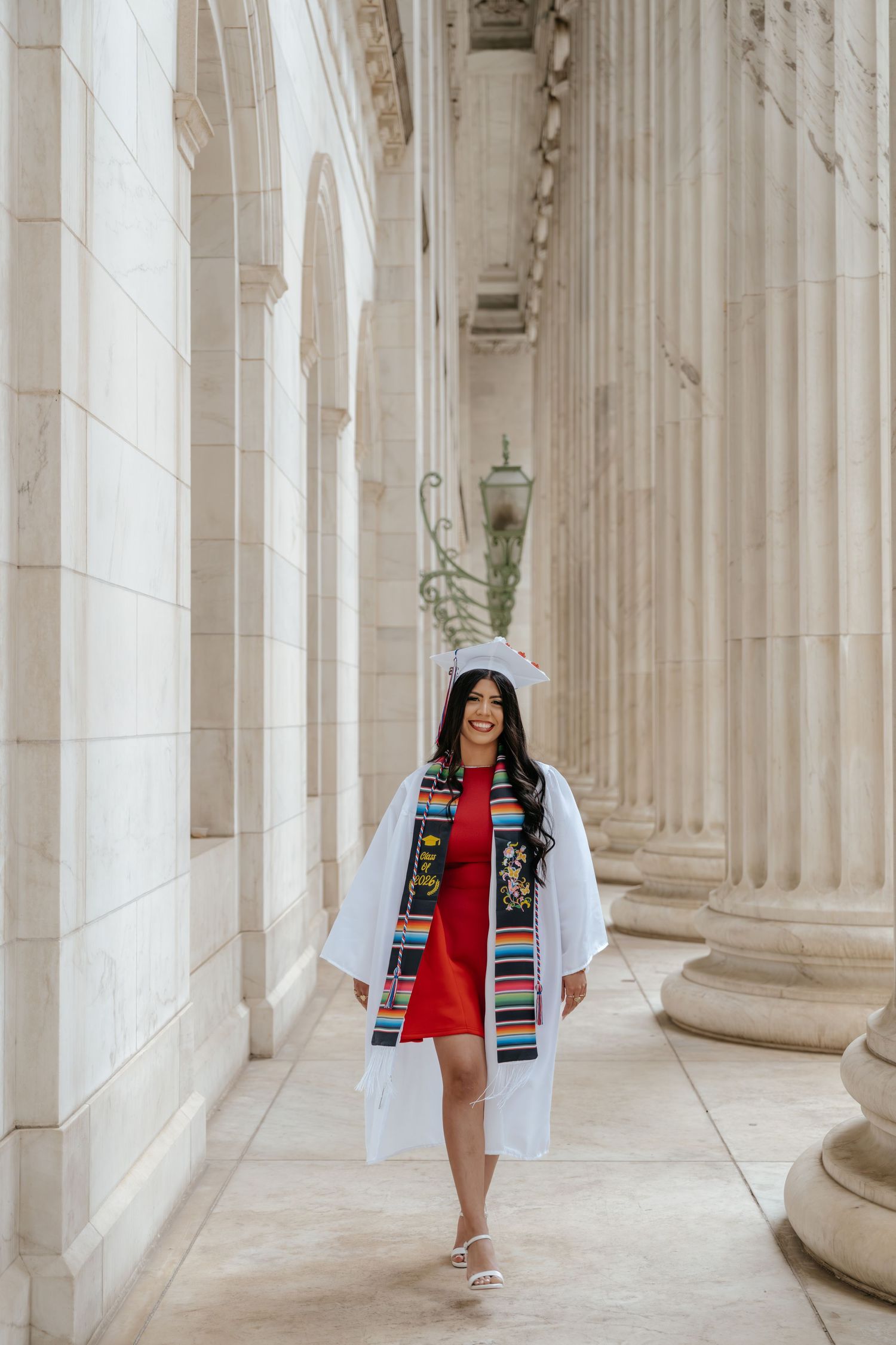 Graduate in white gown and colorful serape stole wearing red dress walks confidently between tall stone columns.
