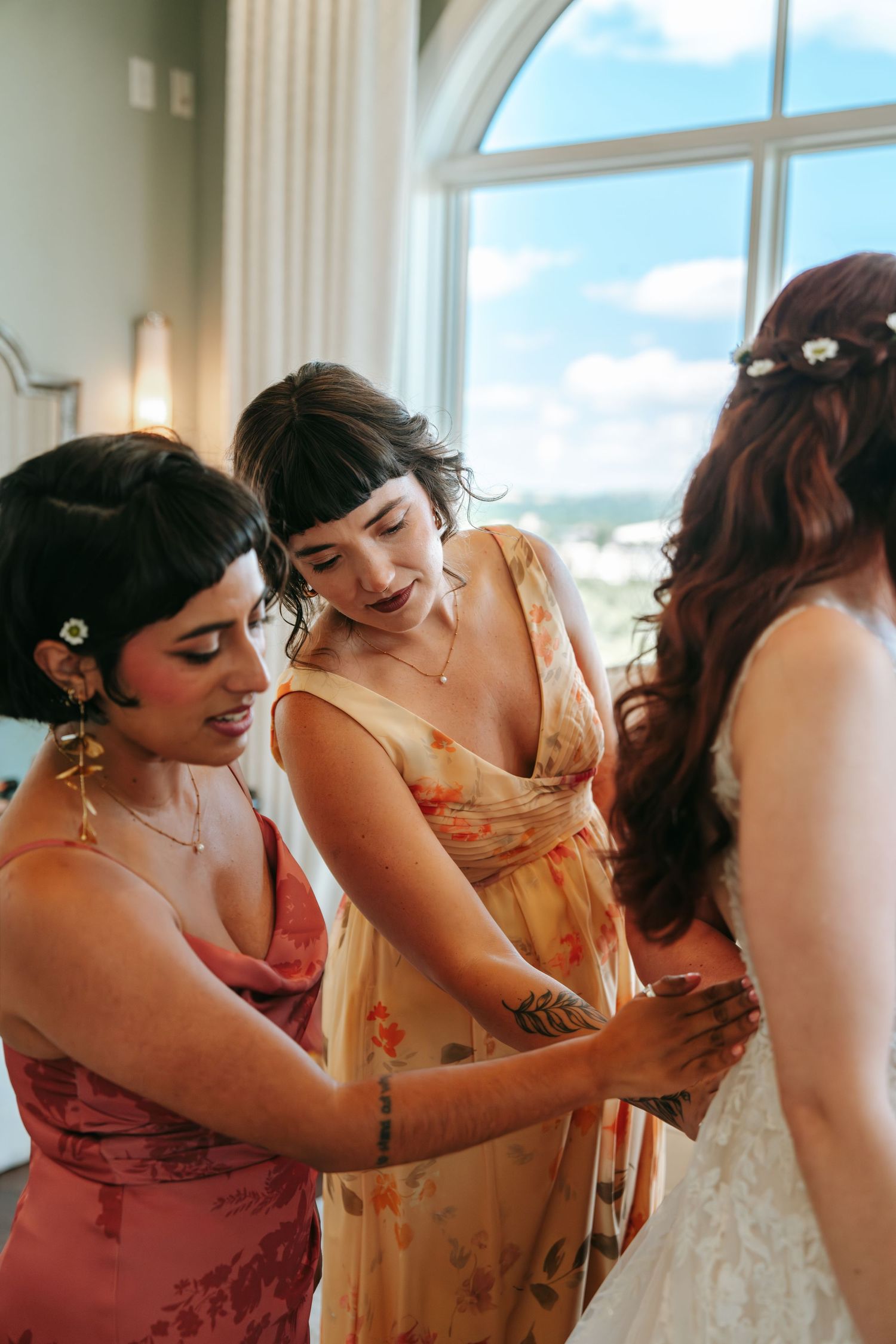 Two bridesmaids in colorful dresses help a bride with her white lace wedding gown near a bright arched window.