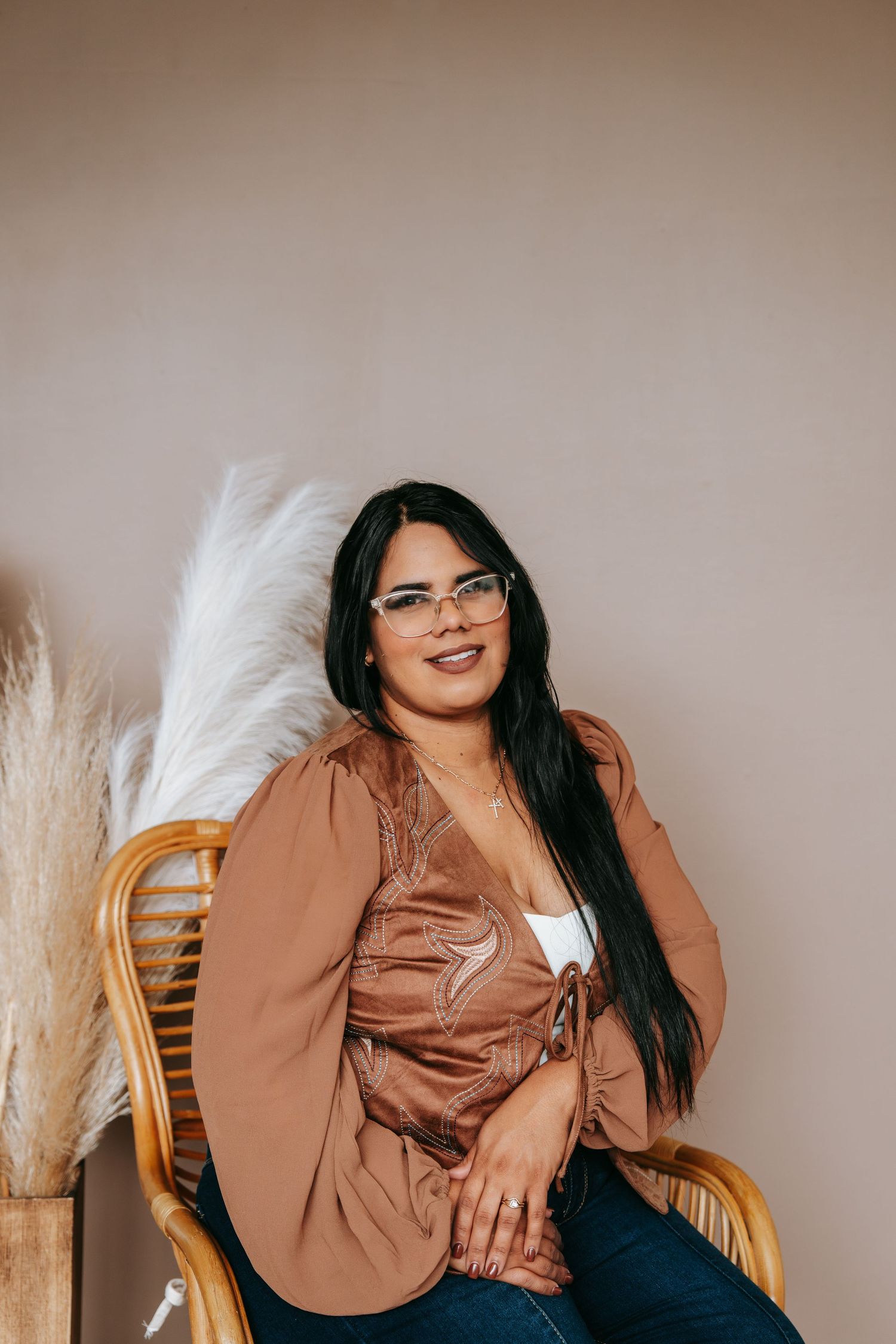 A woman with long dark hair wearing glasses and a brown blouse sits relaxed in a rattan chair.