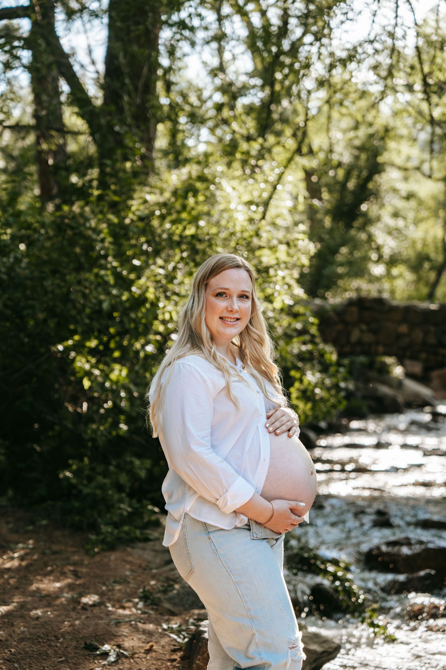 Smiling pregnant woman in white shirt cradles baby bump near a forest stream.