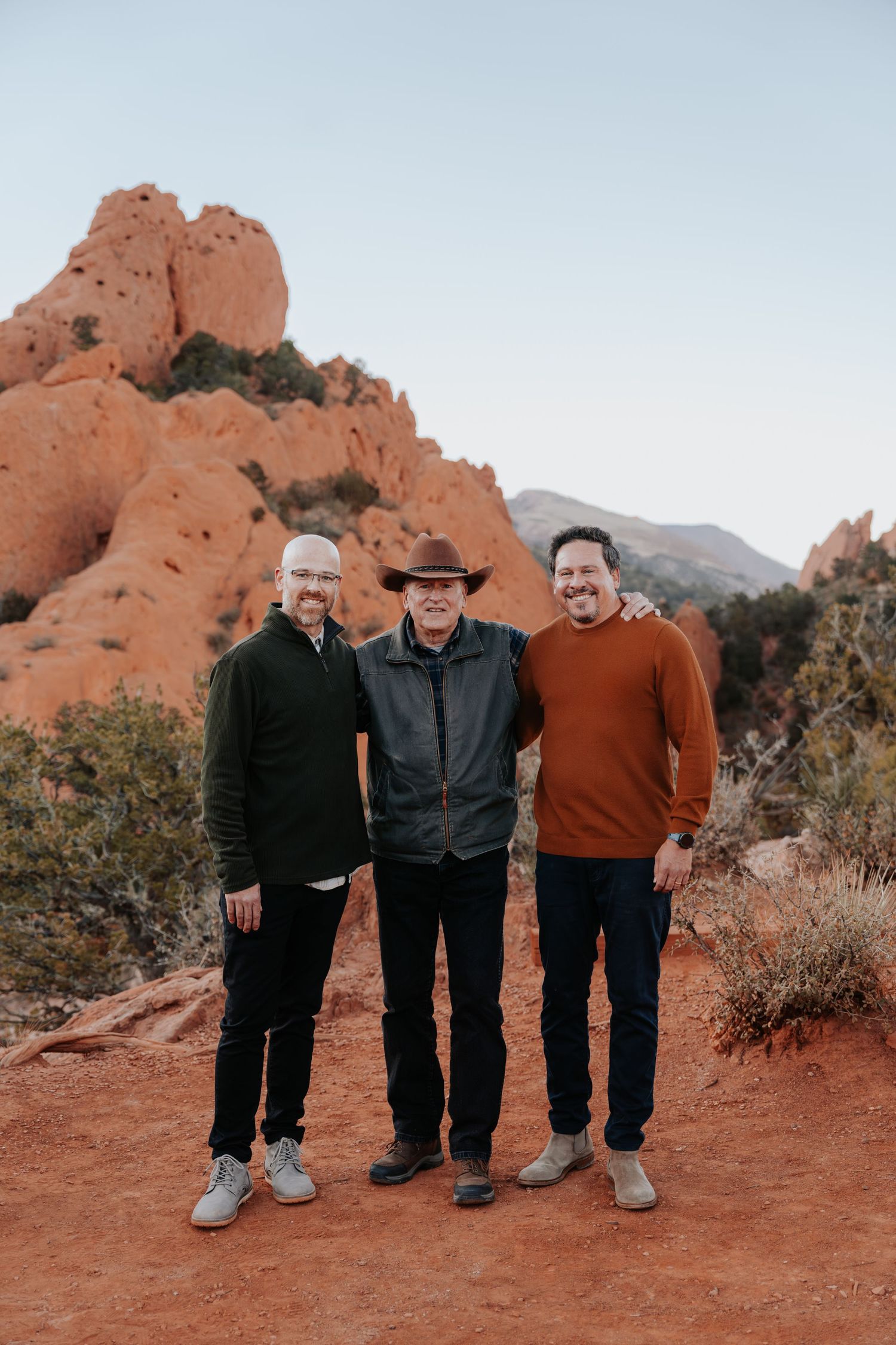 Three men smiling together at Garden of the Gods with red rock formations in Colorado Springs.