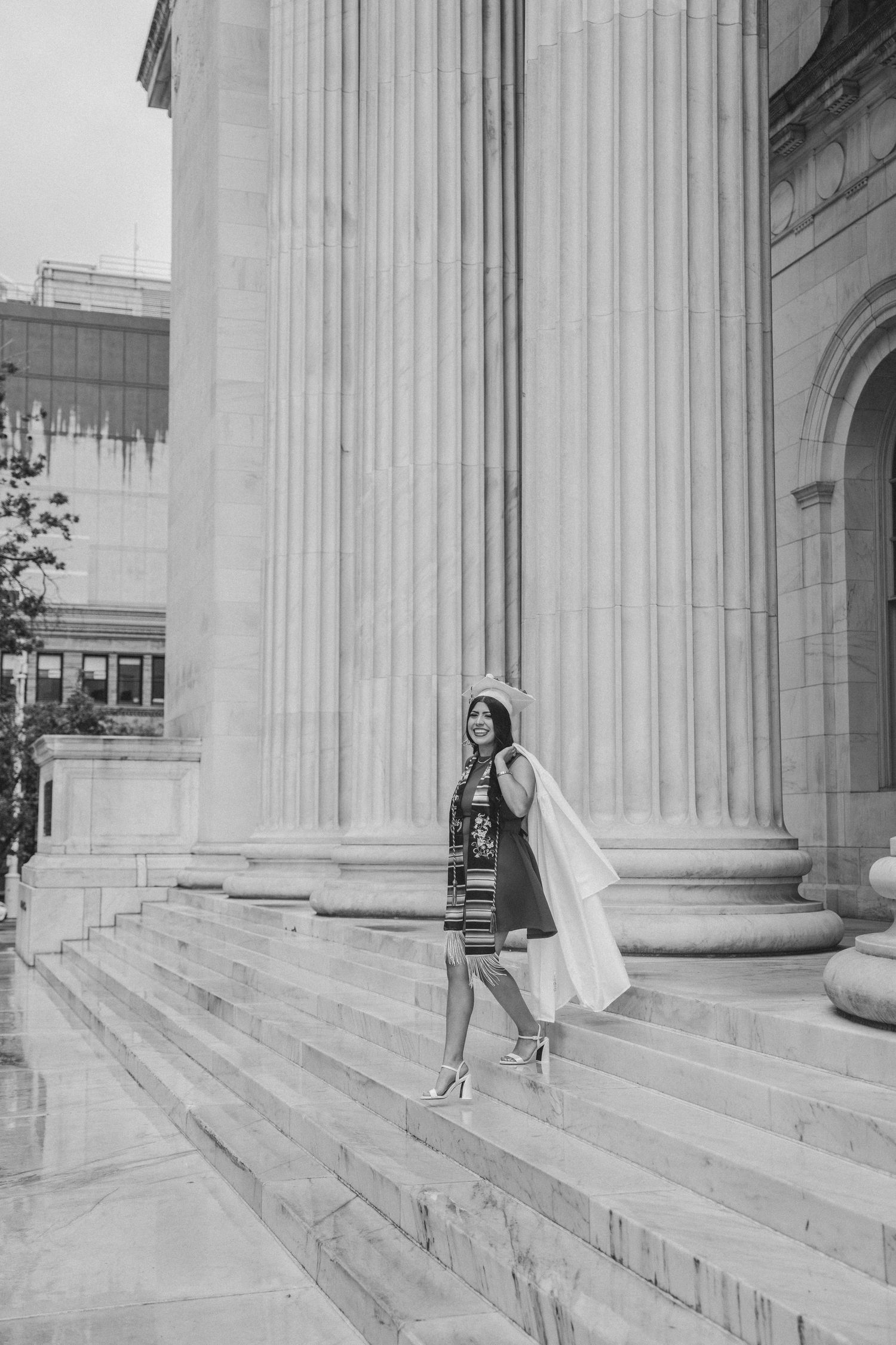 Black and white photo of a graduate in cap and gown descending stone steps beside massive classical columns.